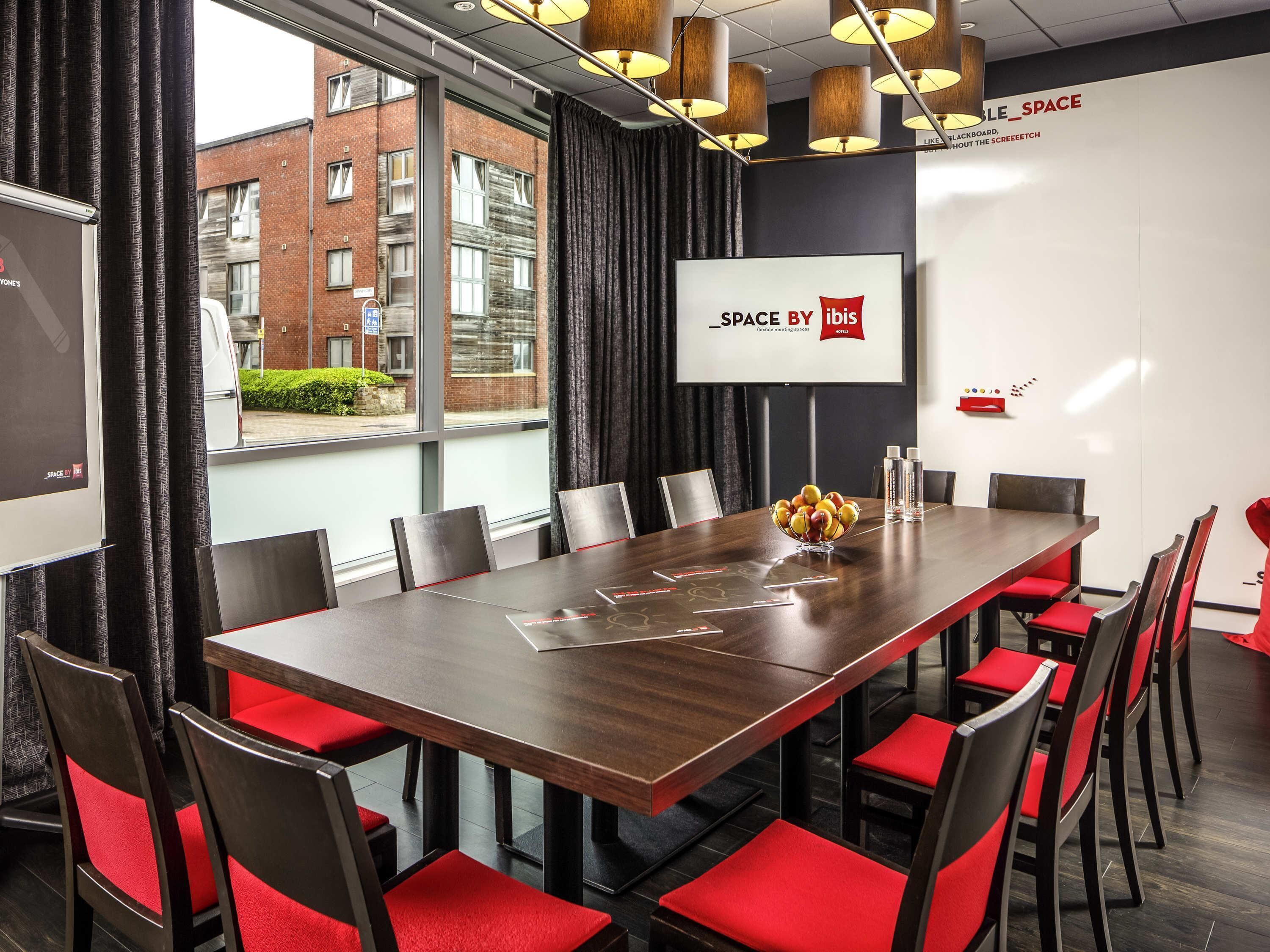 Un salón de conferencias con una larga mesa, sillas rojas, una pantalla de proyector y vistas a un edificio.