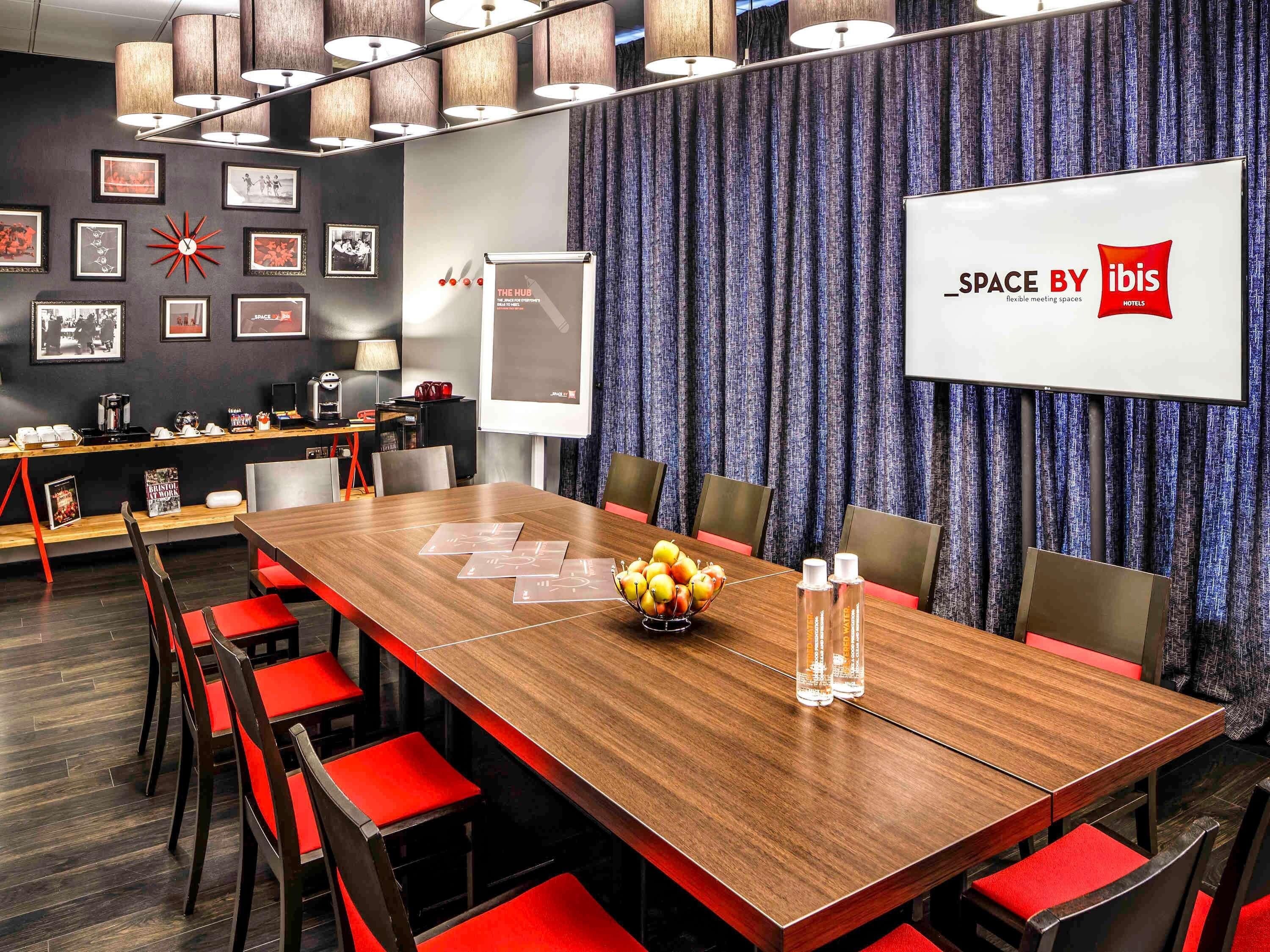 A conference room with a wooden table, red chairs, a projector screen, and a banner reading 'SPACE BY ibis Hotels'.