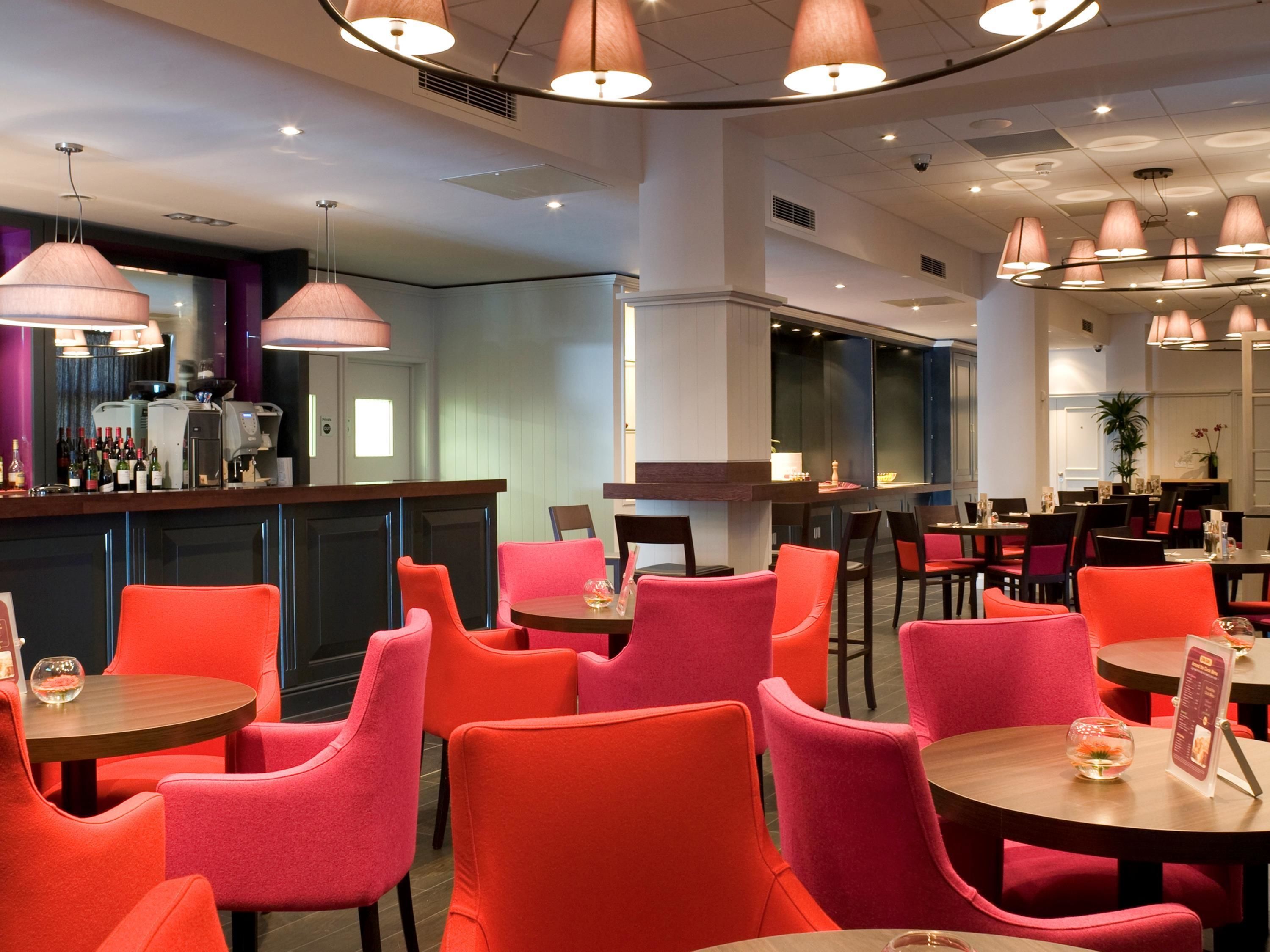 A modern restaurant with round tables and bright orange chairs, featuring a bar area with a coffee machine and wine bottles.