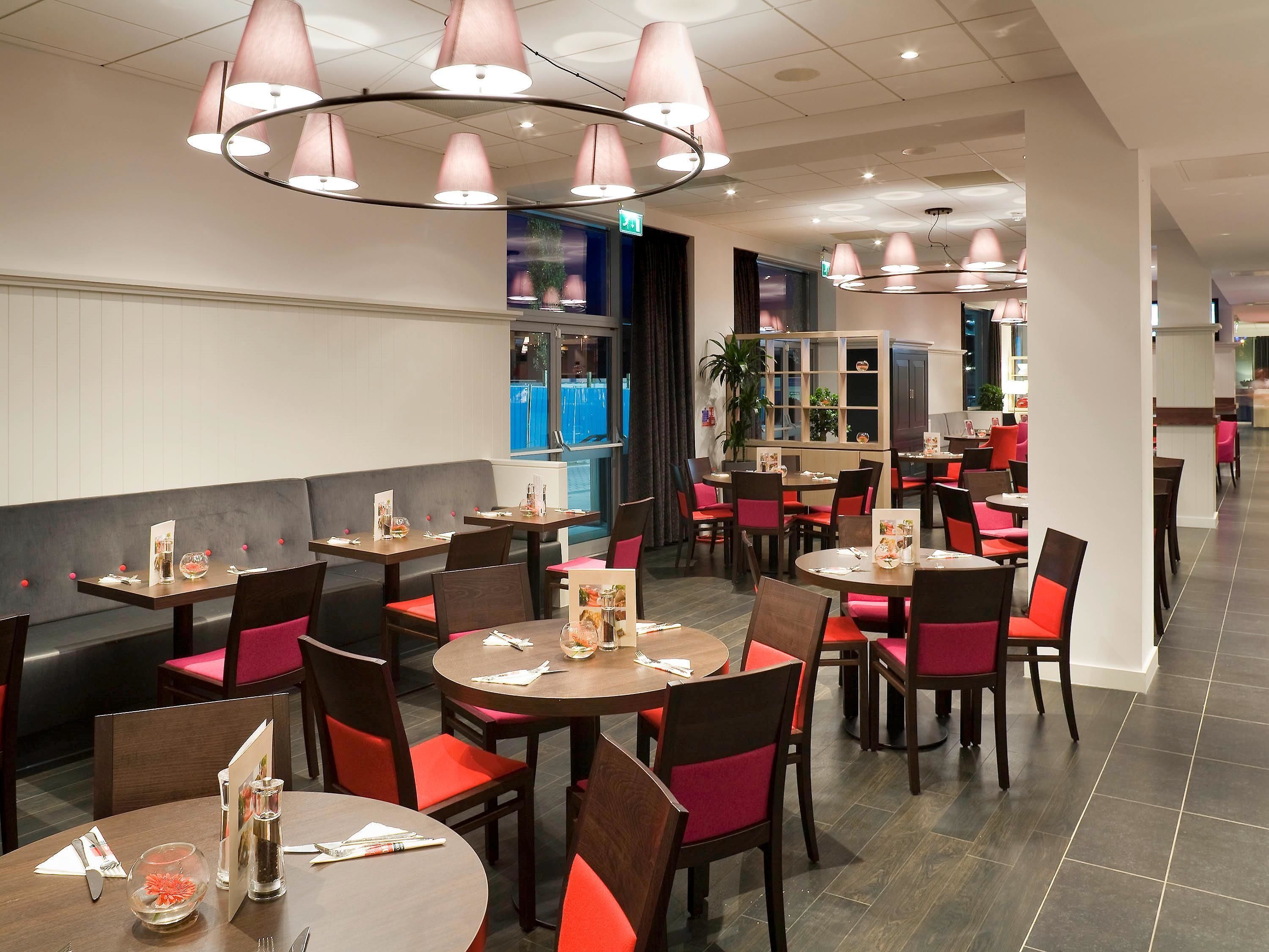 Un restaurante con mesas redondas y sillas rojas acolchadas, con iluminación colgante y vistas al exterior.
