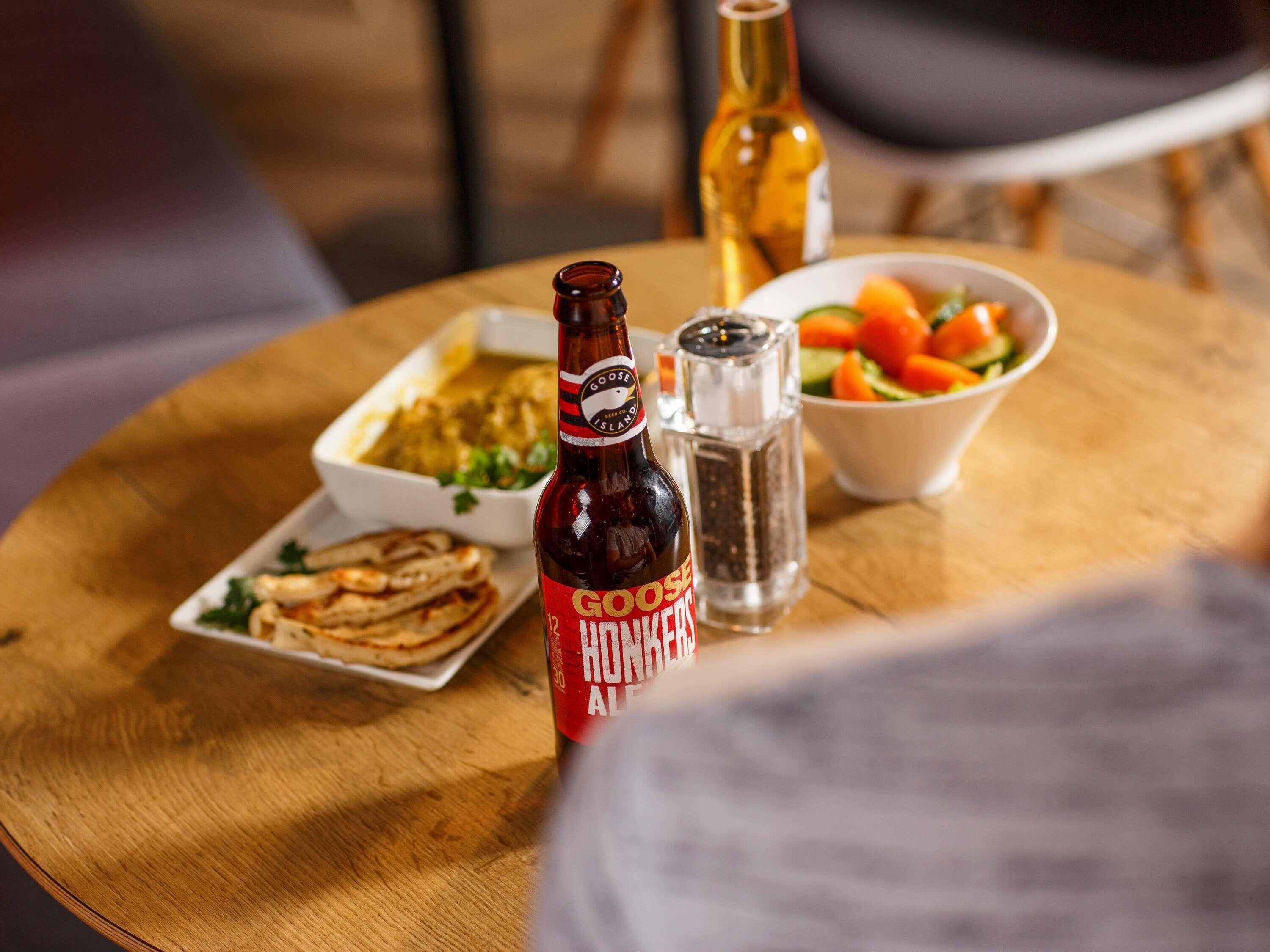 Una mesa de madera con una botella de Goose Island Hot Ales, un tazón de ensalada, un plato de comida y un salero.