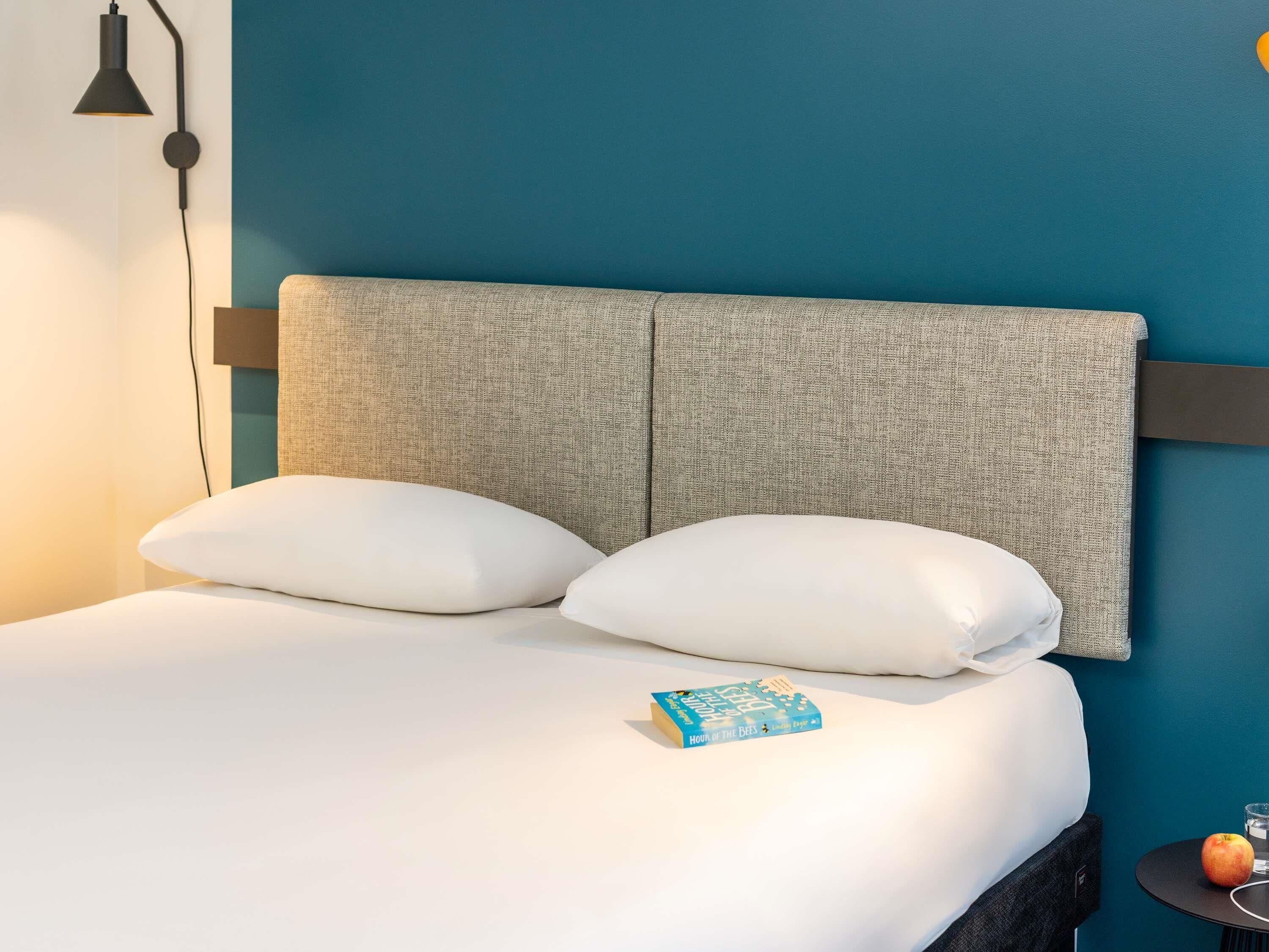 A bed with a headboard, pillows, a book, a glass of water, and an apple on a small table.