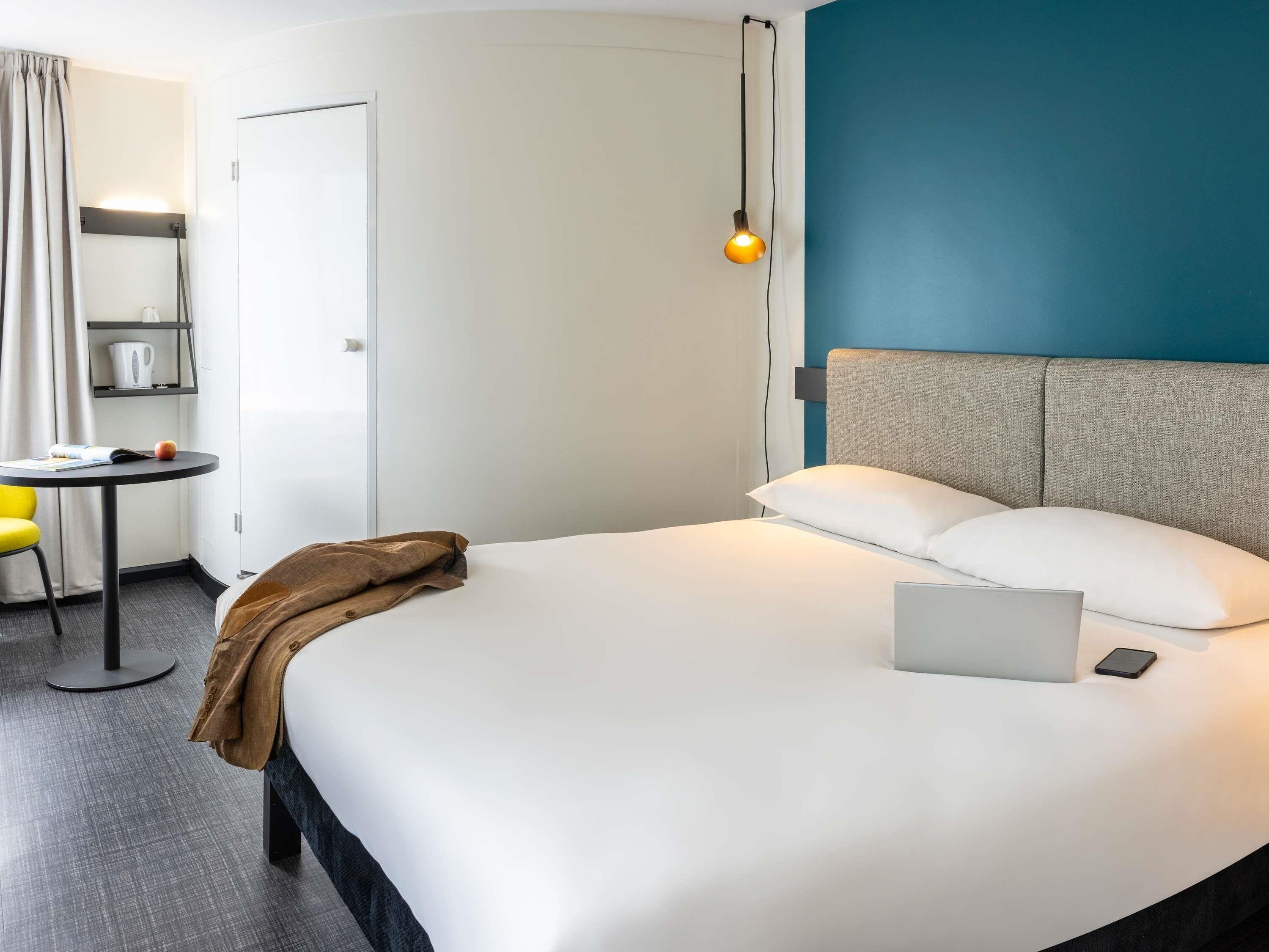 A modern hotel room with a neatly made bed, a laptop on the bed, and a small table with a kettle and a book.