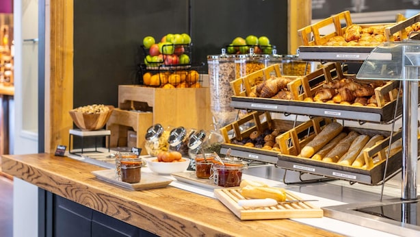 Mostrador de panadería con diversas opciones horneadas, incluyendo cruasanes, muffins y pasteles, junto con frascos de mermelada y un bol de frutas.