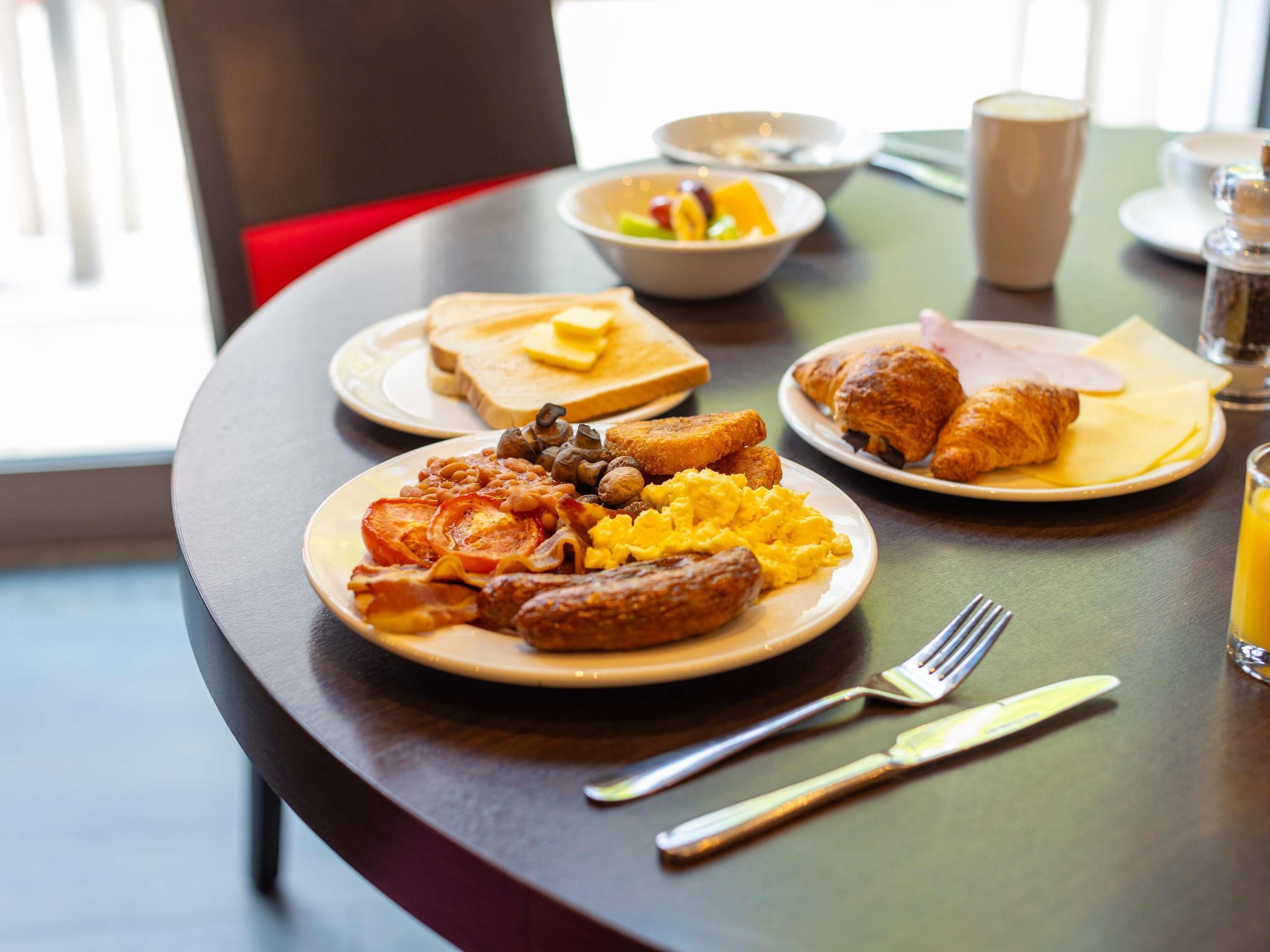 A breakfast setting with plates of eggs, sausages, and toast, a croissant, and a glass of orange juice on a table.