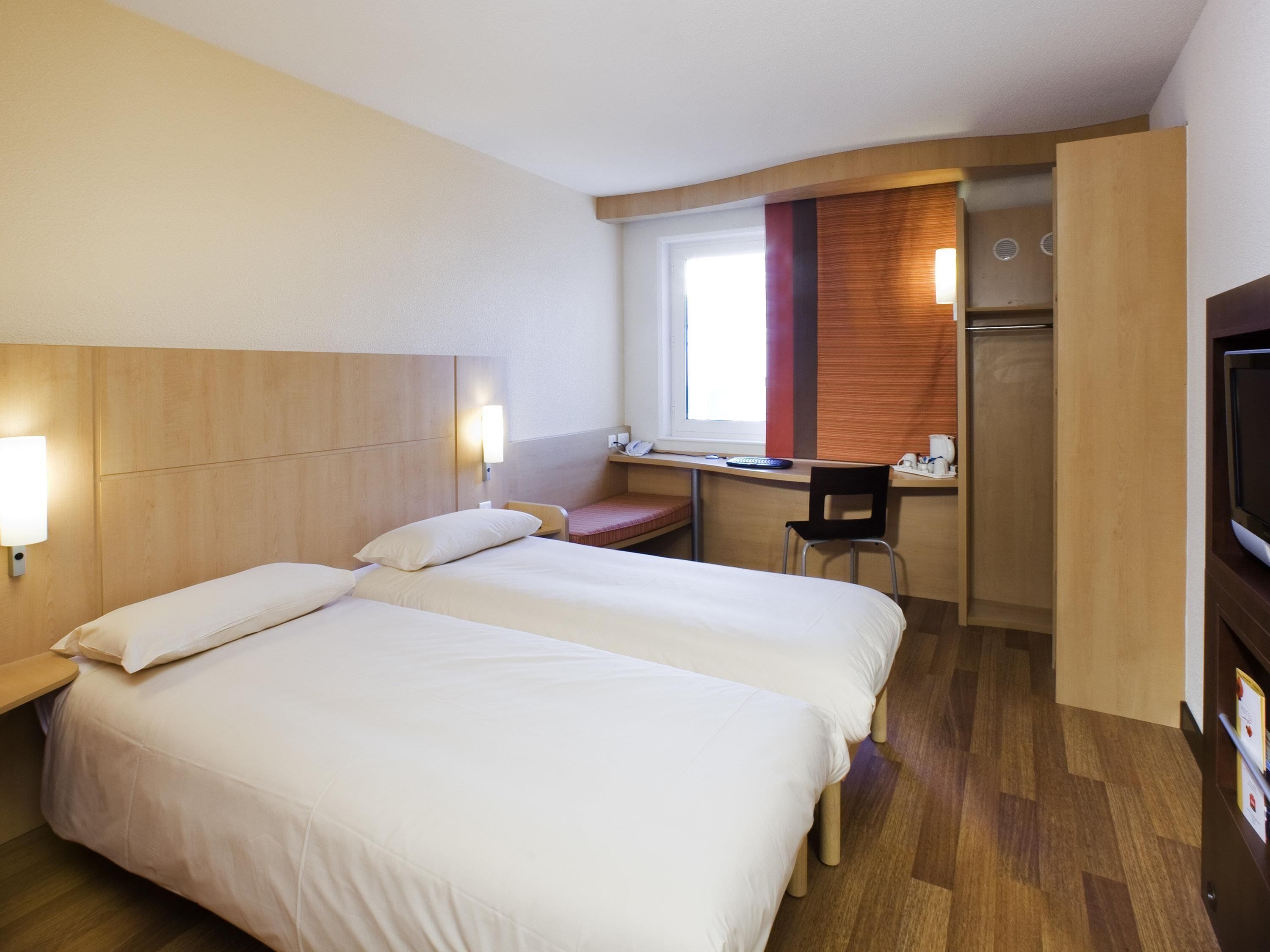 A hotel room with two beds, a desk, a chair, a TV, and a window.