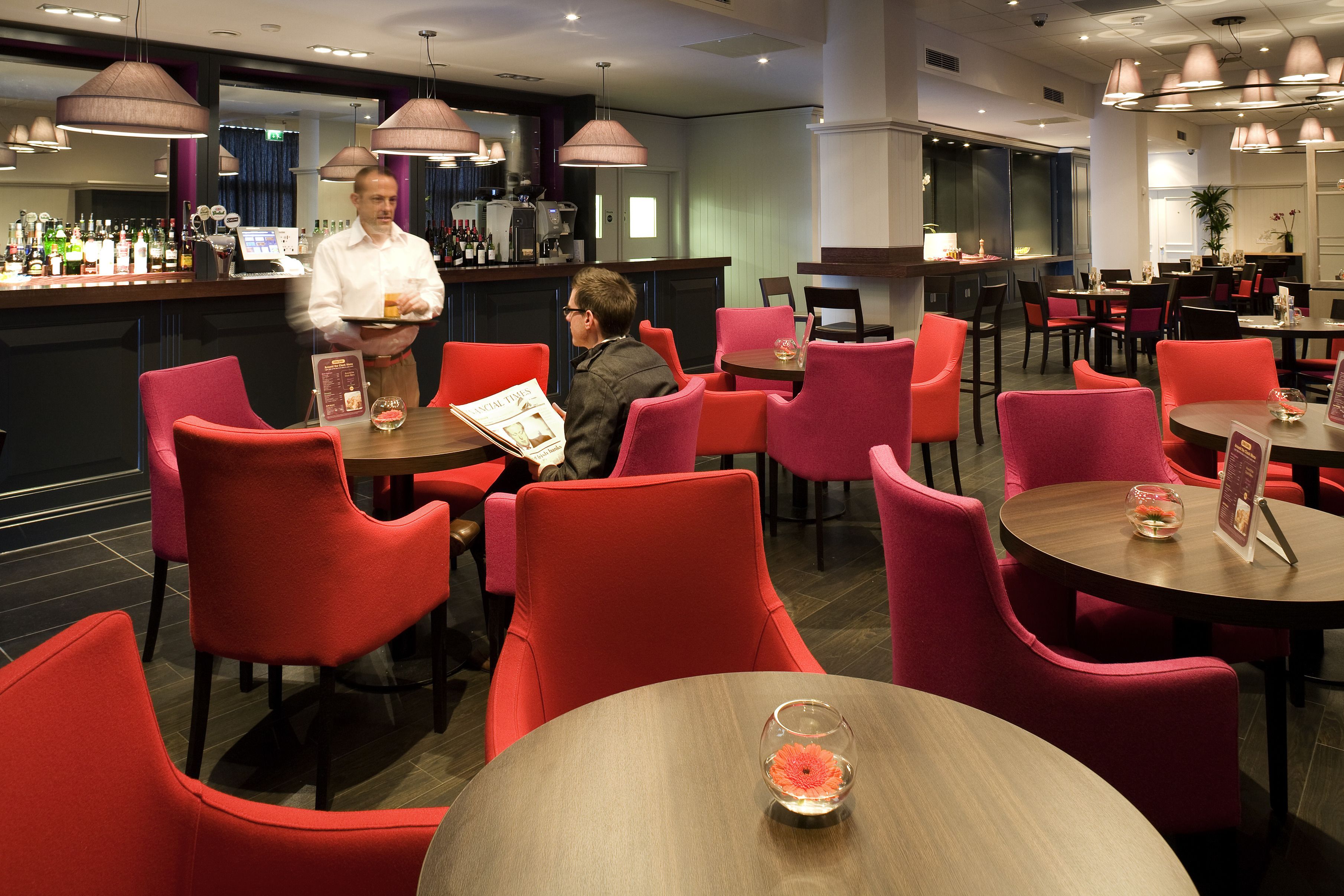 A modern restaurant with red chairs and wooden tables, a bar area with a bartender, and a menu board.