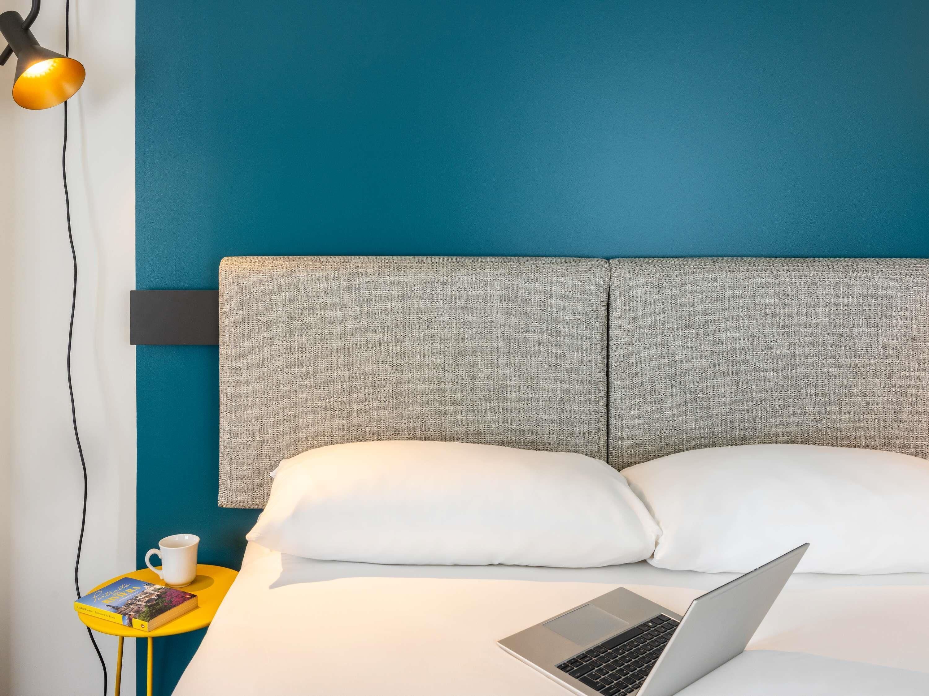Una cama con almohadas blancas, una laptop sobre la cama, una mesita con un libro y una taza, y una lámpara fijada a la pared.