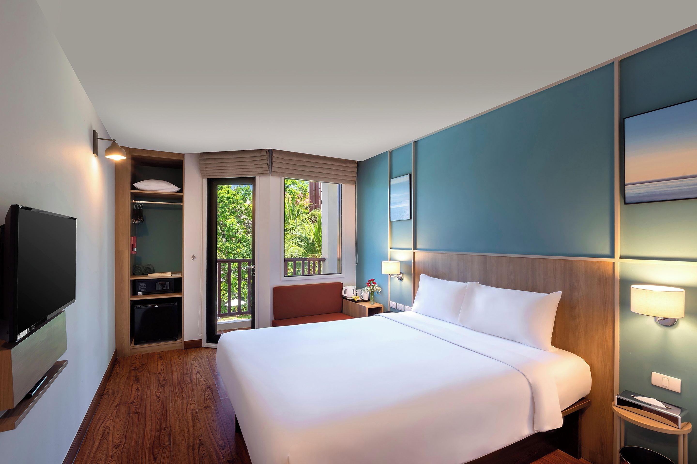 A hotel room with a bed, a television, a desk with a lamp, and a view of greenery outside.