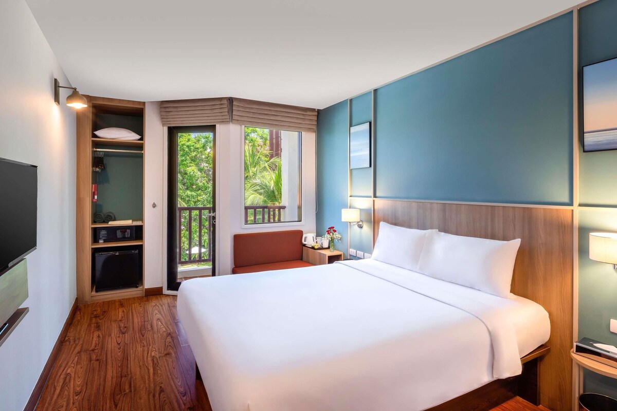A hotel room with a large bed, a TV, a wardrobe, and a balcony with a view of greenery.