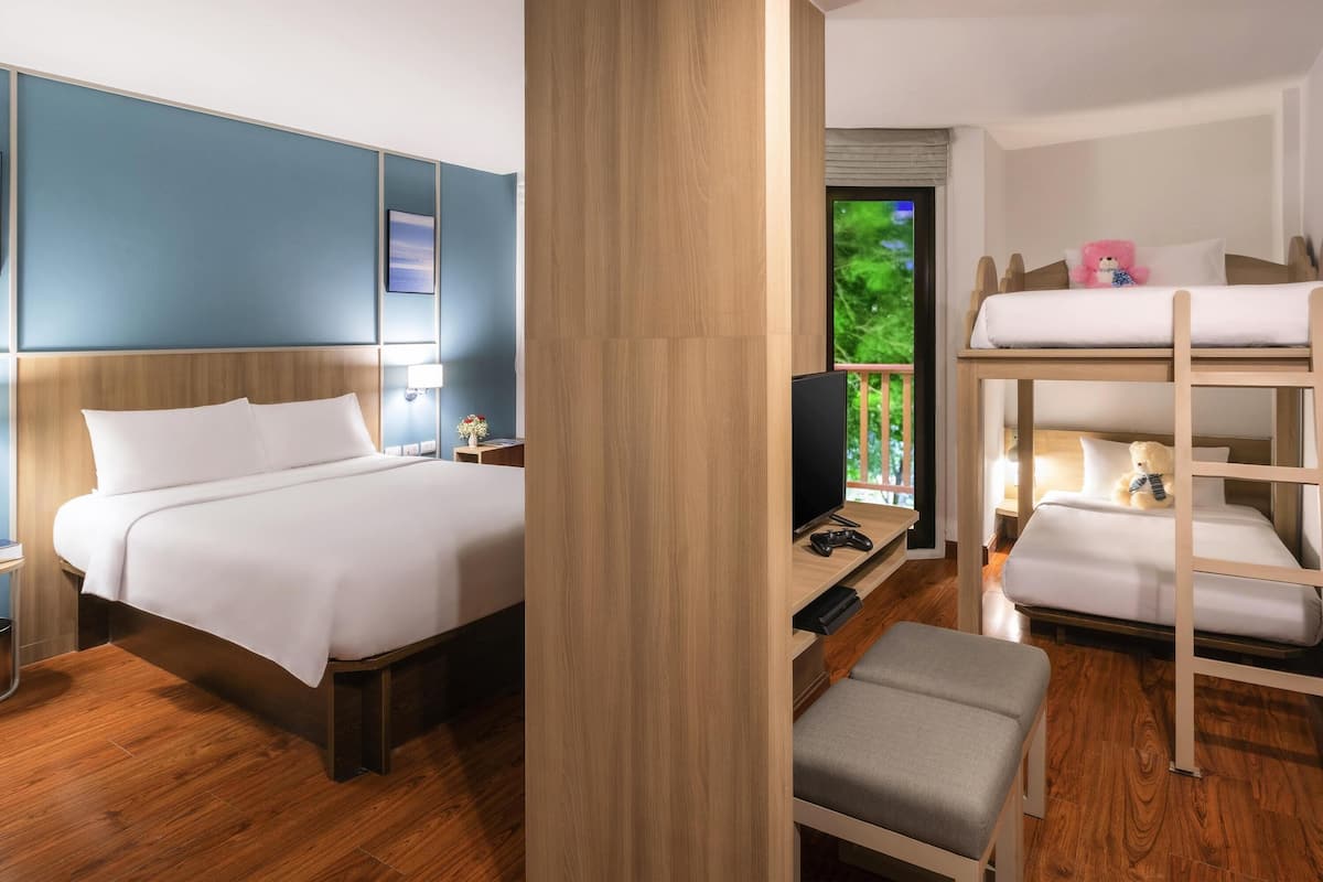 A hotel room with a single bed, a desk with a computer, a TV, a bunk bed, and a window with a view of greenery.