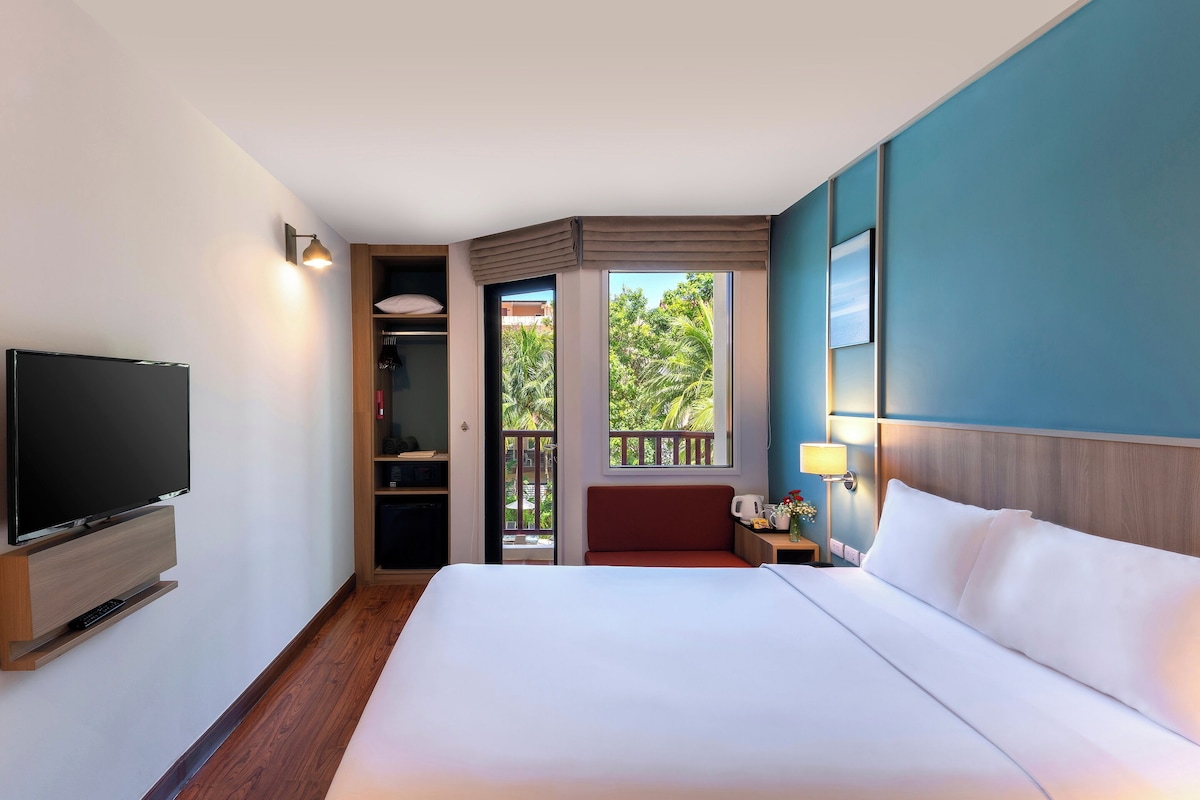 A hotel room with a bed, TV, wardrobe, and a balcony with a view of greenery.