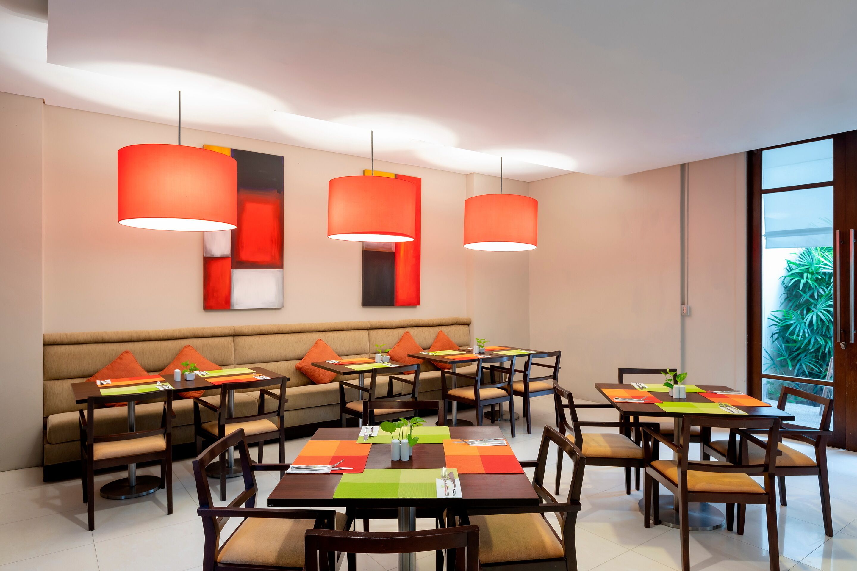 Un comedor moderno con varias mesas y sillas, lámparas colgantes naranjas y un ventanal con vistas a zonas verdes.