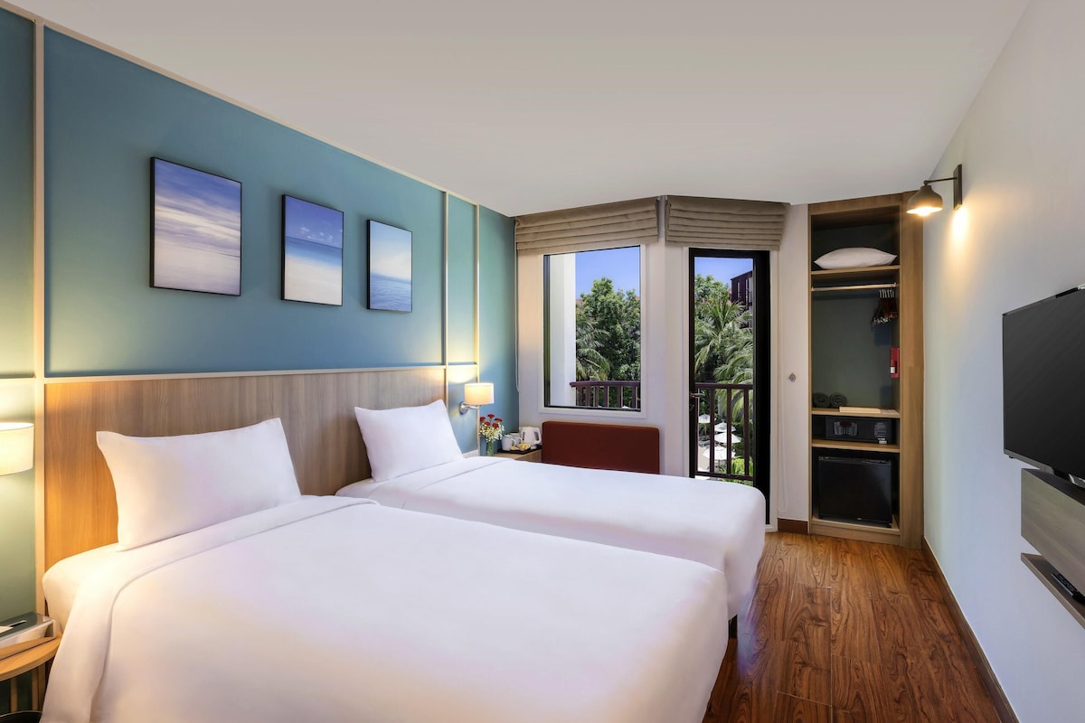 A hotel room with two beds, a TV, a wardrobe, and a balcony with a view of trees.