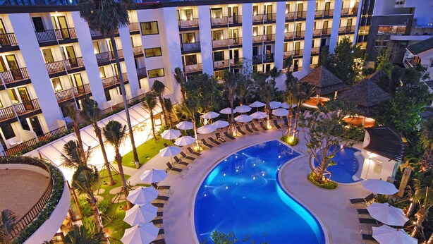 Un hotel con una amplia piscina rodeada de sillas de descanso y sombrillas, varios pisos con balcones y un exterior bien iluminado.