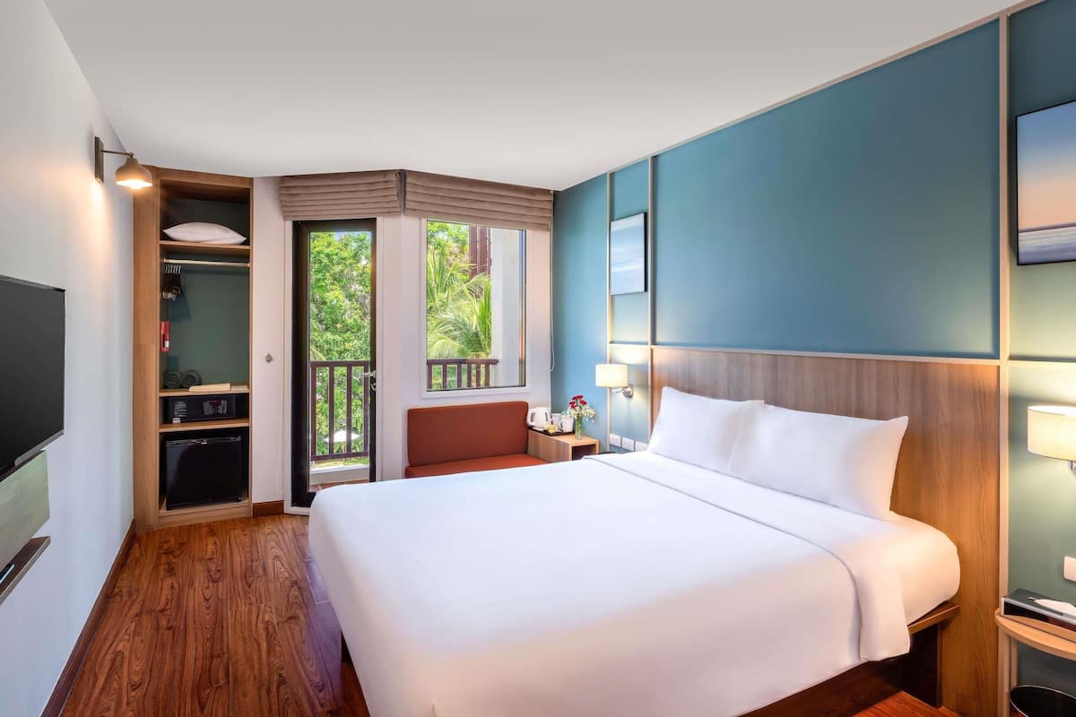 A hotel room with a large bed, a TV, a wardrobe, and a balcony with a view of greenery.
