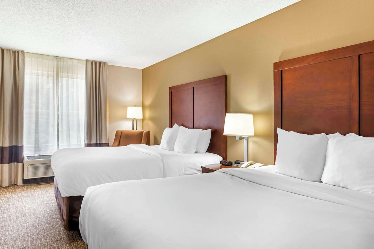 A hotel room with two beds, a wooden headboard, a bedside table with a lamp, and a window with curtains.