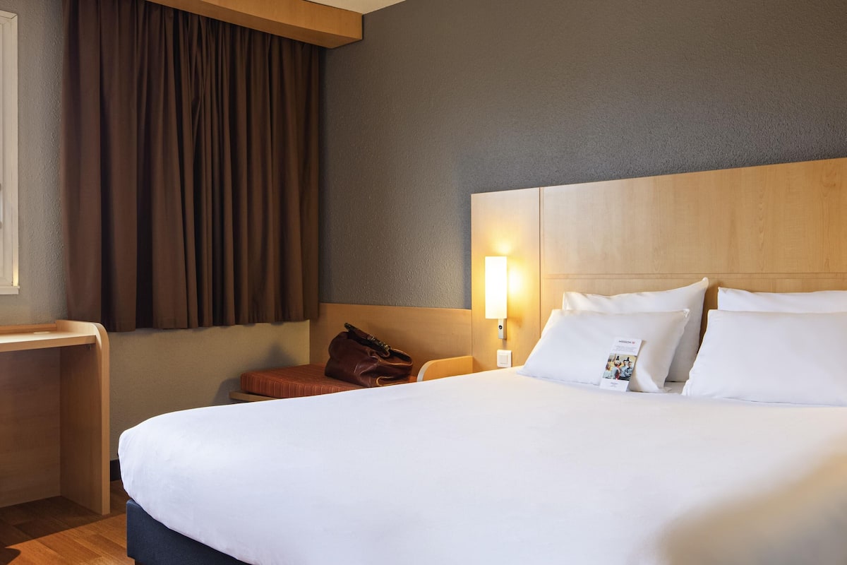 A hotel room with a large bed, a bedside table, a lamp, and a window with curtains.