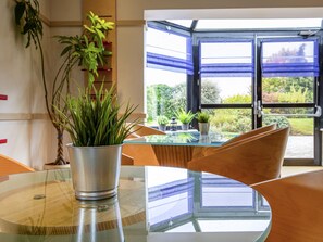 A modern dining area with a glass table, metal plant pot, and a view through a large window with blue blinds.
