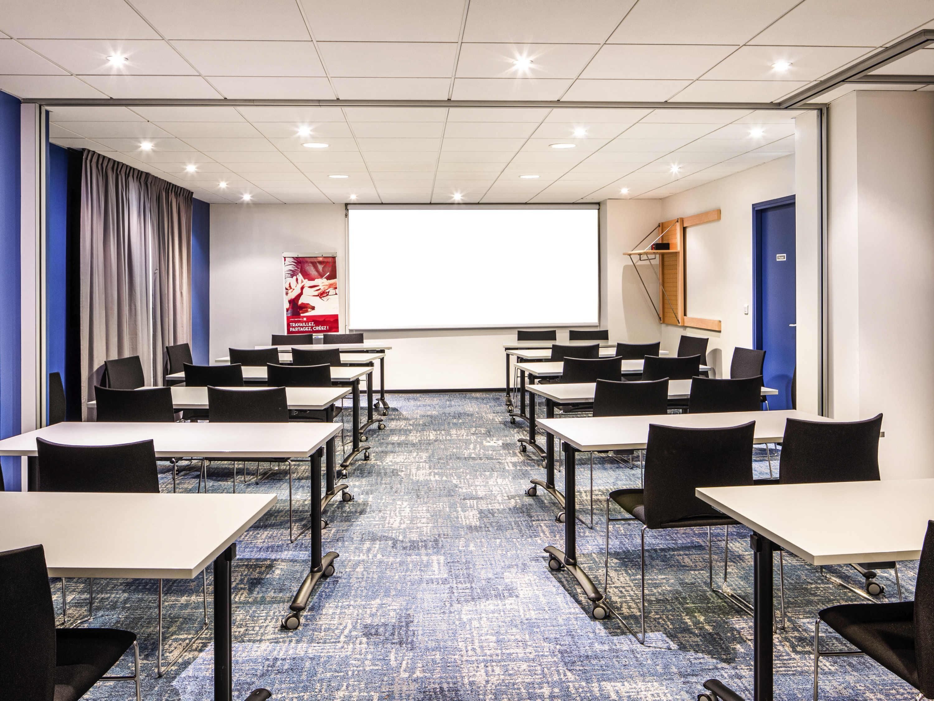 A conference room with tables, chairs, a projector screen, and a carpeted floor.