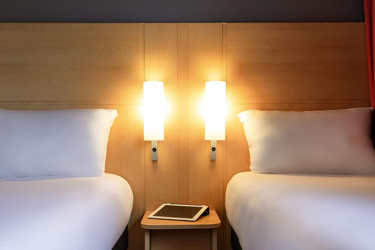 A hotel room with two beds, each with a white pillow and a bedside table with a tablet.