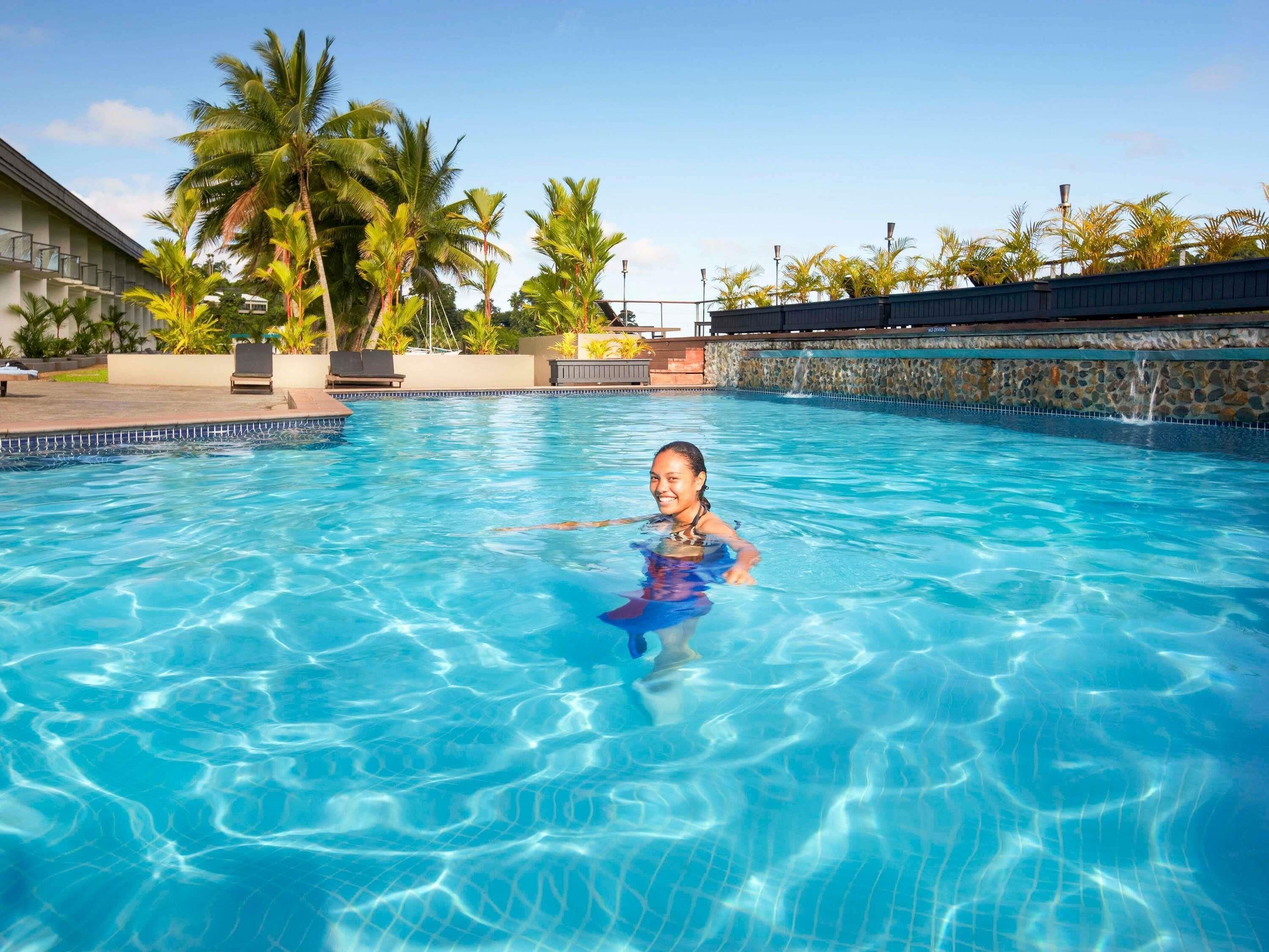 Une piscine à l’eau d’un bleu cristallin, entourée de palmiers et d’un mur de pierre.