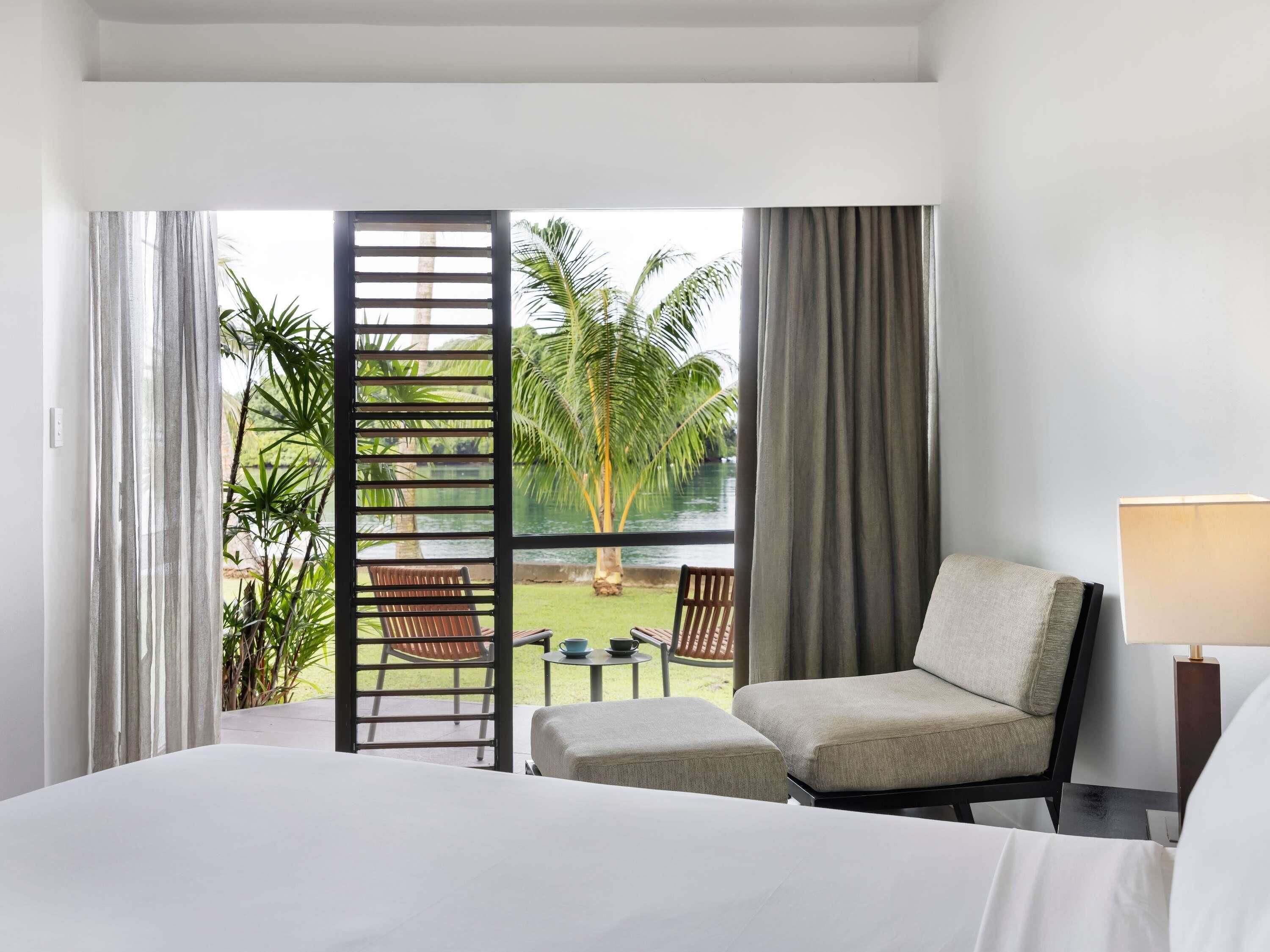 A hotel room with a bed, a chair, a table, and a view of a pool and palm trees.