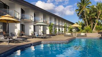 A hotel with a swimming pool, sun loungers, umbrellas, and palm trees.