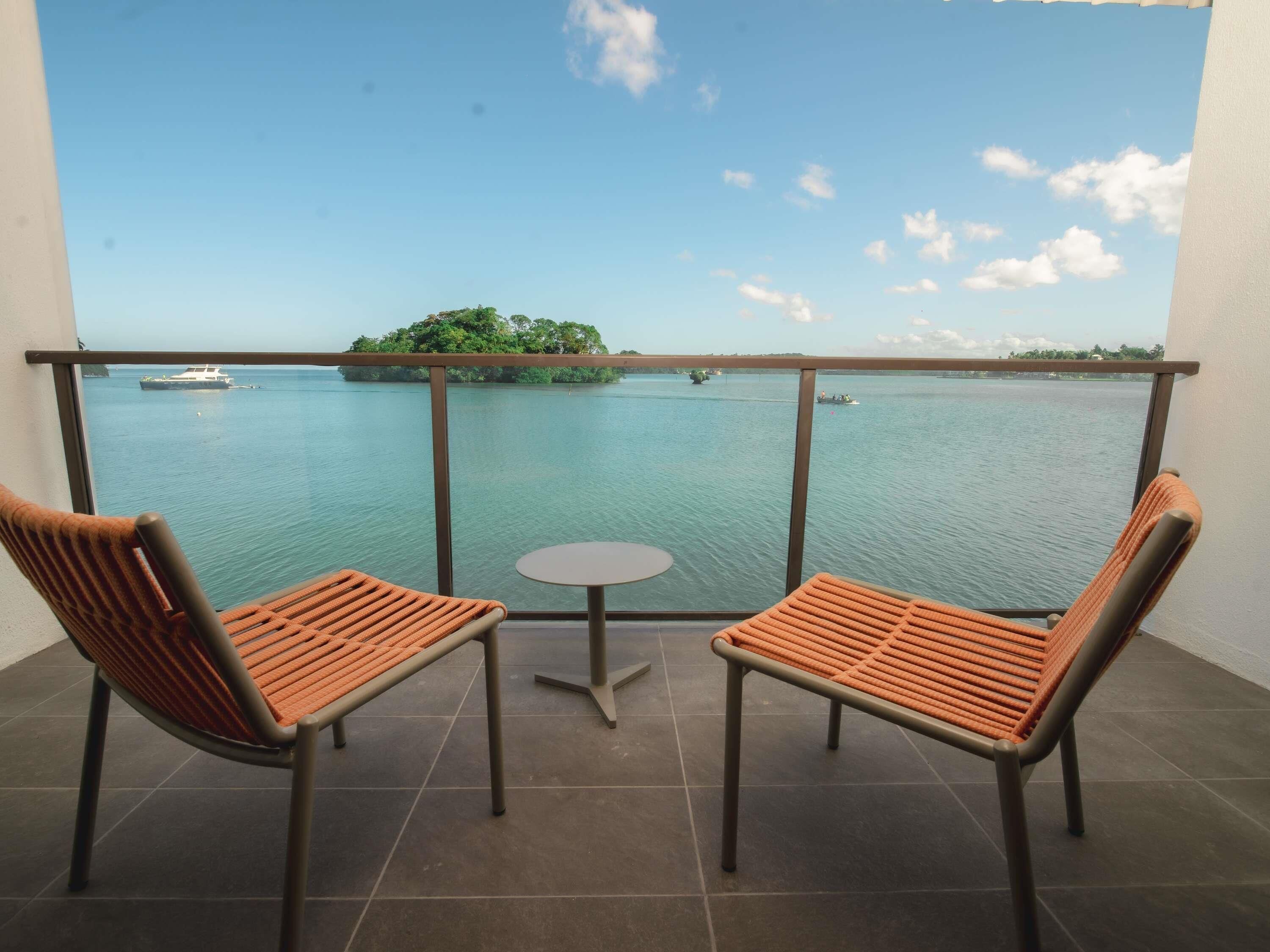 A balcony with two chairs and a small table, overlooking a calm body of water with boats and an island.