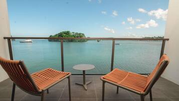 A balcony with two chairs and a small table, overlooking a calm body of water with boats and an island.