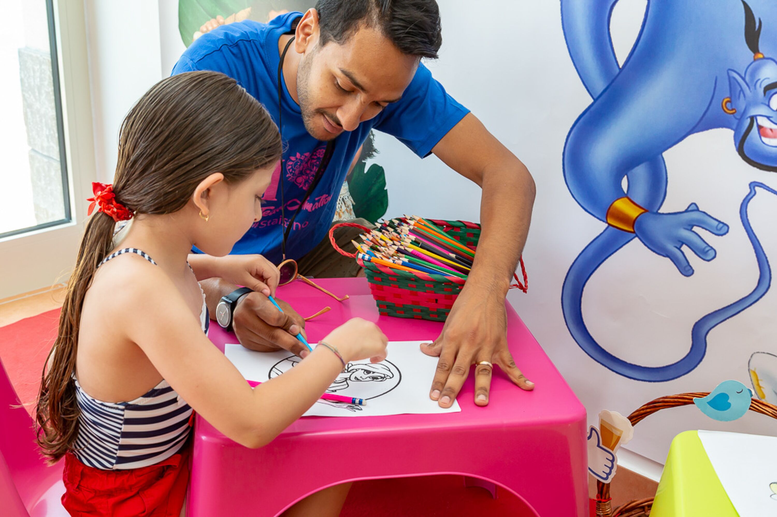 A child drawing at a table with a pink tablecloth, a basket of colored pencils, and a large cartoon character on the wall.