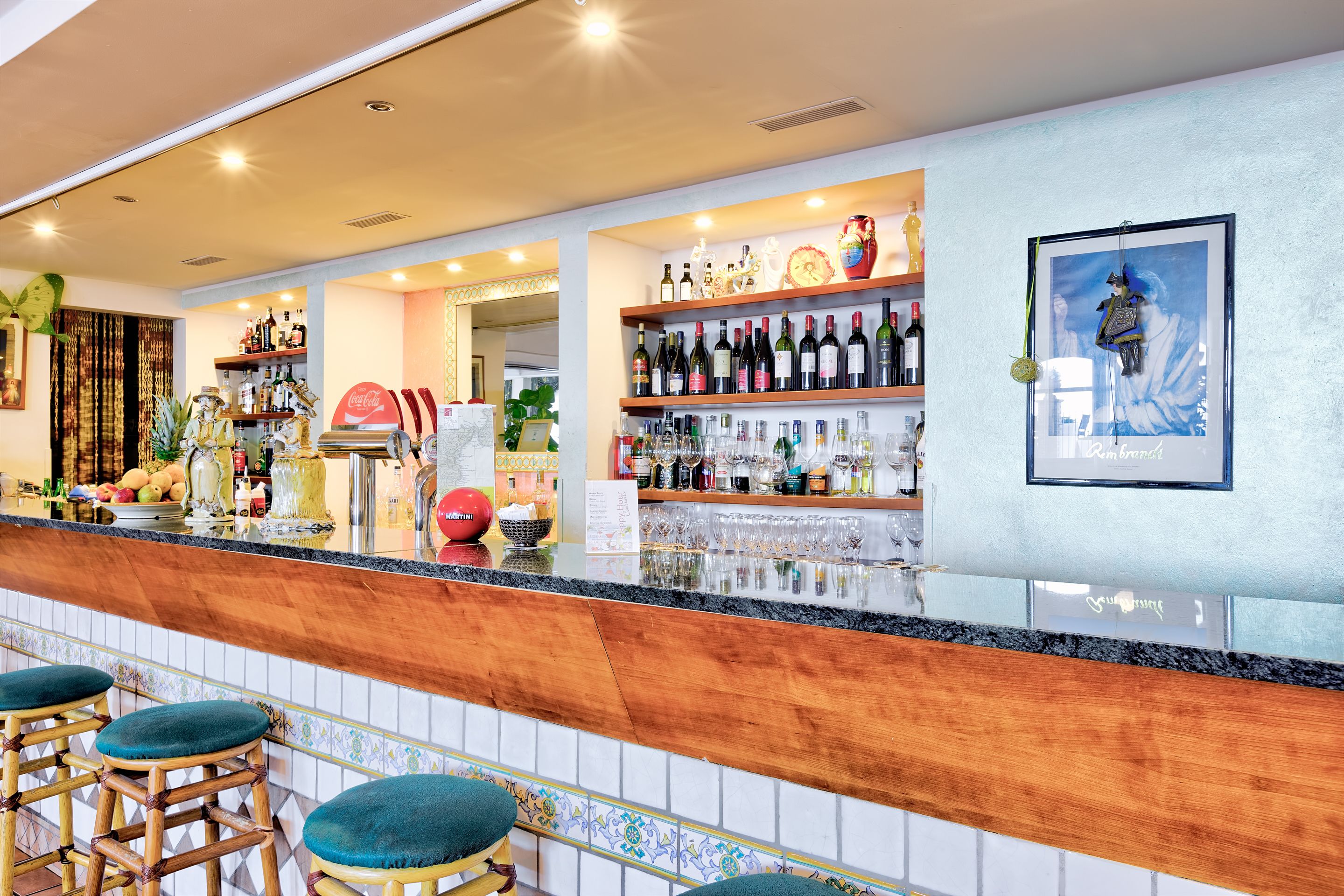 A bar area with a wooden counter, bar stools, shelves with bottles, and a framed picture on the wall.