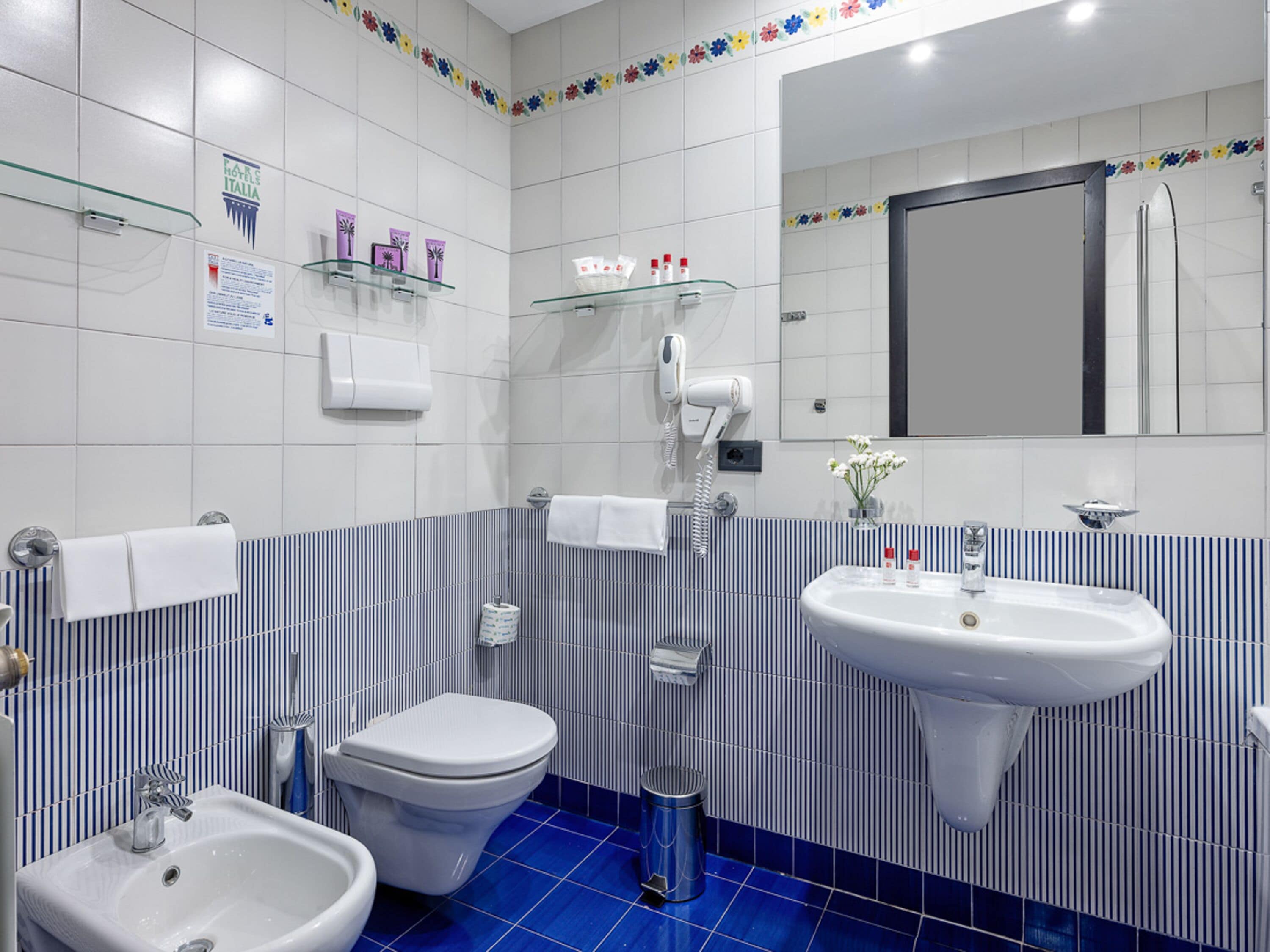 Une salle de bain moderne équipée de toilettes, d’un bidet et d’un lavabo. On y trouve des étagères avec des articles de toilette et un miroir au-dessus du lavabo.