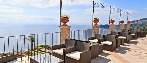 A terrace with wicker furniture and potted plants, overlooking a coastal view.