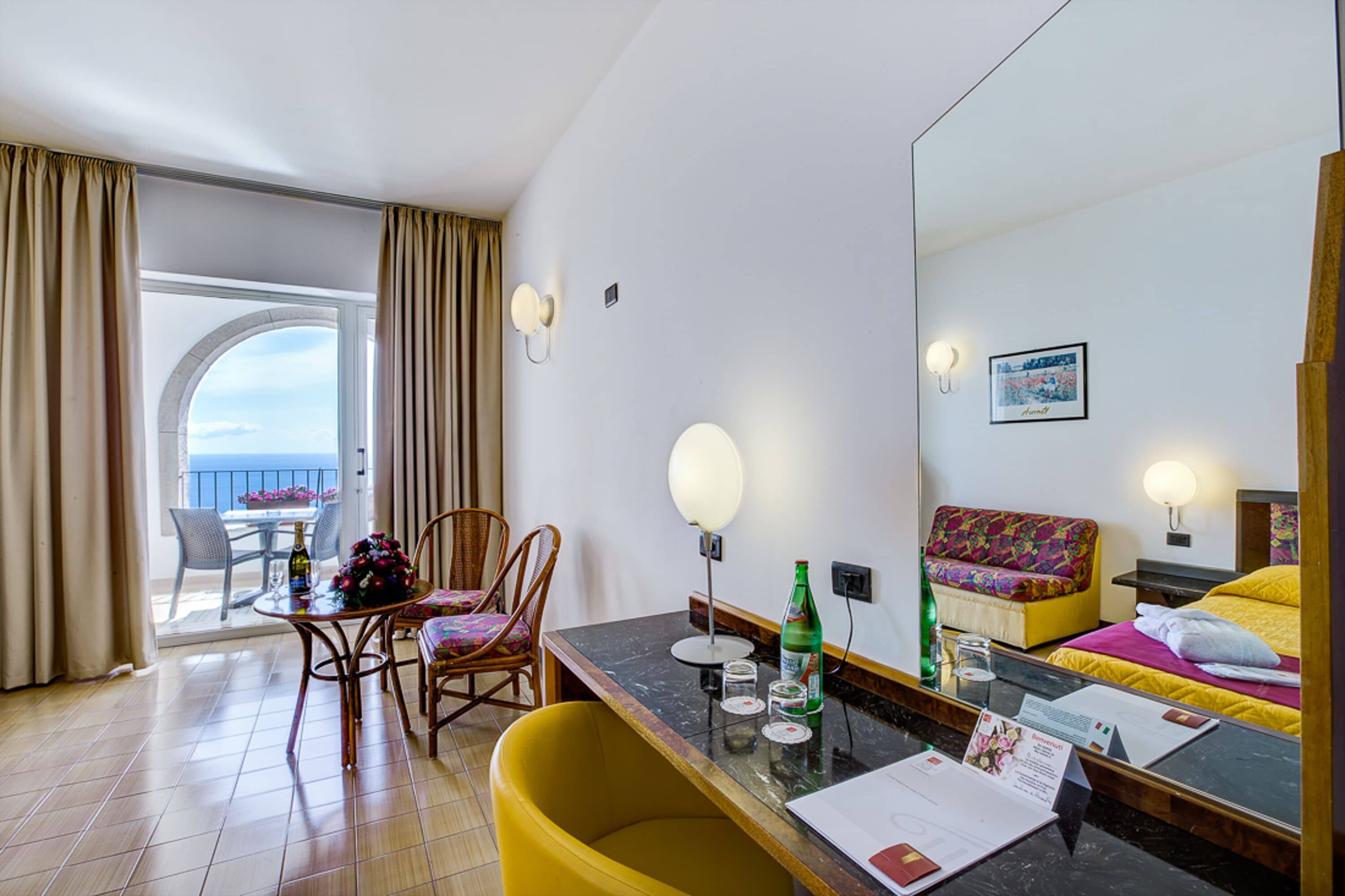 A hotel room with a balcony, a dining area with chairs and a table, a bed with a colorful headboard, and a large mirror reflecting the room.