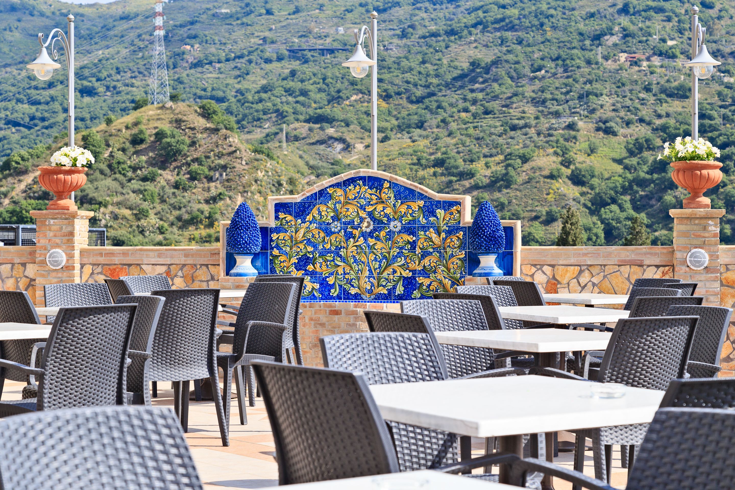 Uma área de refeições ao ar livre com cadeiras e mesas de vime, uma parede decorativa com azulejos e vista para uma paisagem montanhosa.