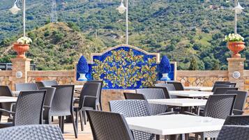 Uma área de refeições ao ar livre com cadeiras e mesas de vime, uma parede decorativa com azulejos e vista para uma paisagem montanhosa.