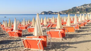 Een strand met rijen oranje ligbedden en beige parasols, een heldere lucht en een heuvelachtig landschap op de achtergrond.