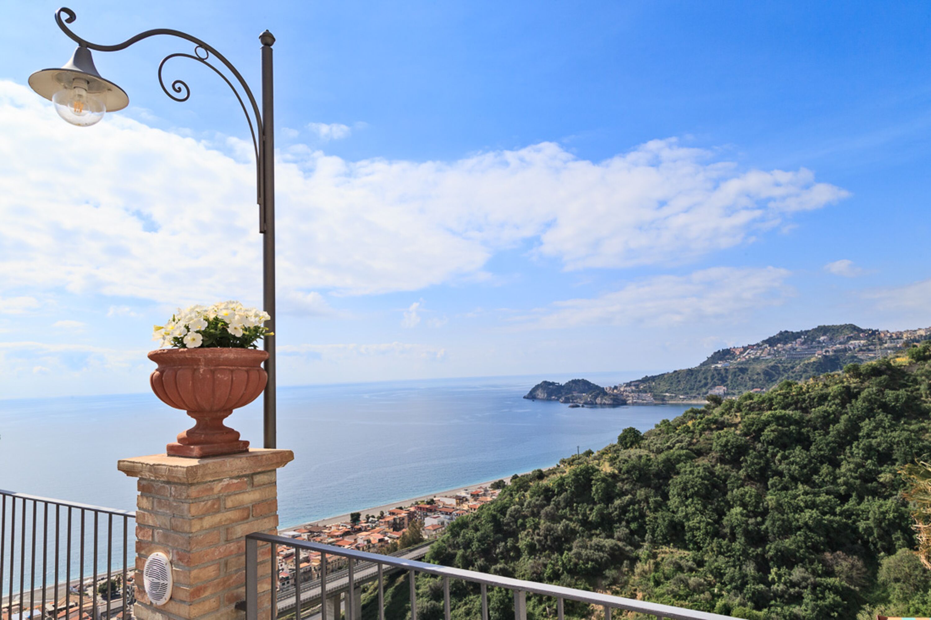 A balcony with a flower pot, overlooking a coastal town and the sea.
