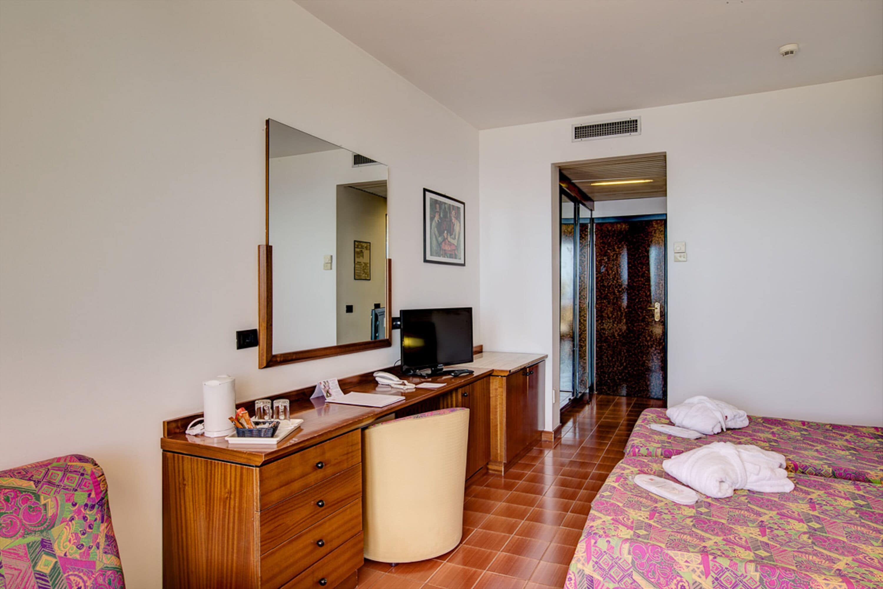 A hotel room with a wooden desk, a television, a bed with a colorful bedspread, and a large mirror.