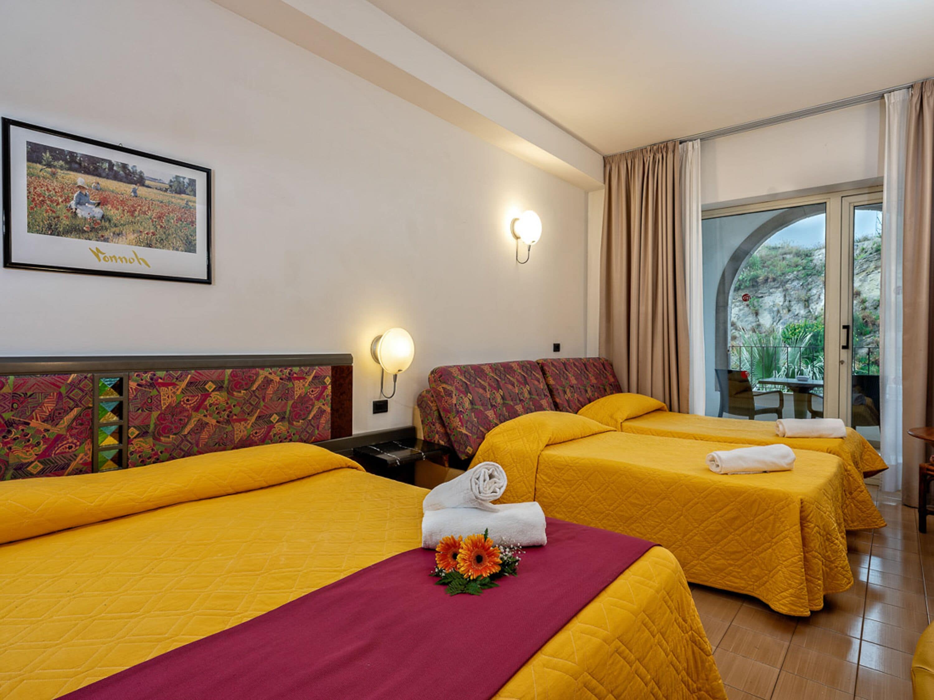 A hotel room with three beds, each with a yellow bedspread and a red accent. There is a framed picture on the wall, a window with curtains, and a balcony with a table and chairs.