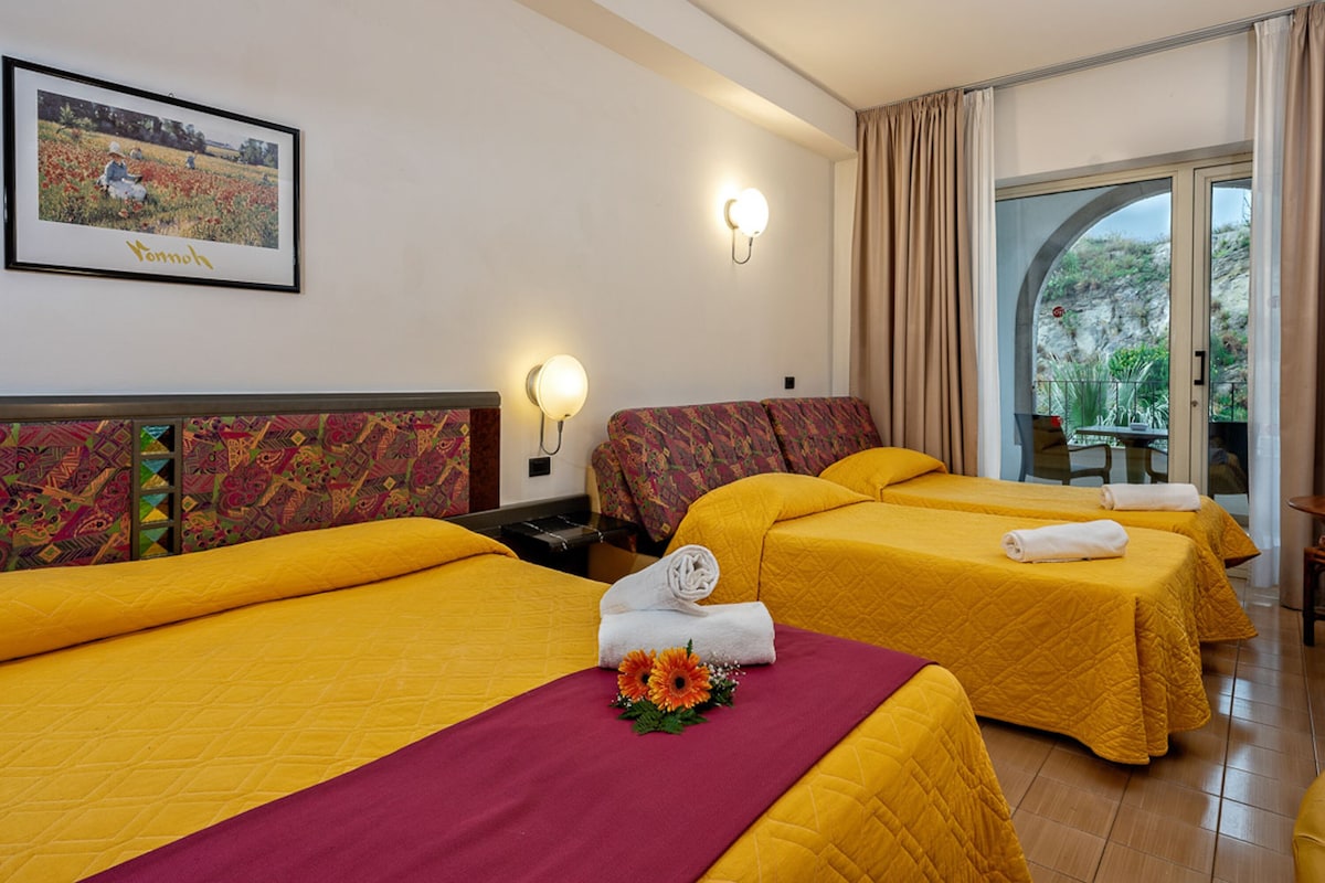 A hotel room with three beds, each with a yellow bedspread and a red accent. There is a framed picture on the wall, a window with curtains, and a balcony with a table and chairs.