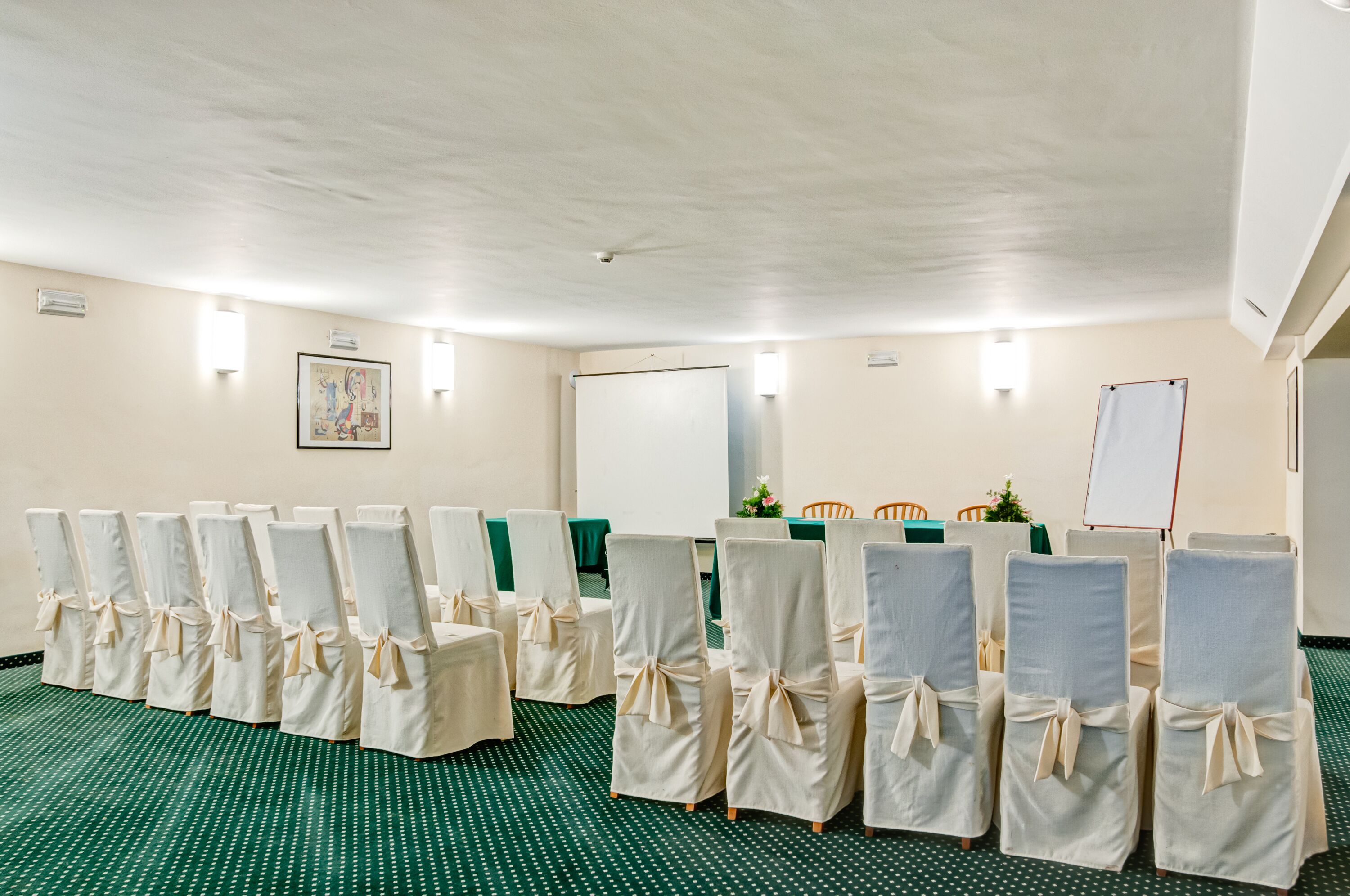 Een vergaderruimte met rijen stoelen bekleed met witte stof, een whiteboard en een projectiescherm.