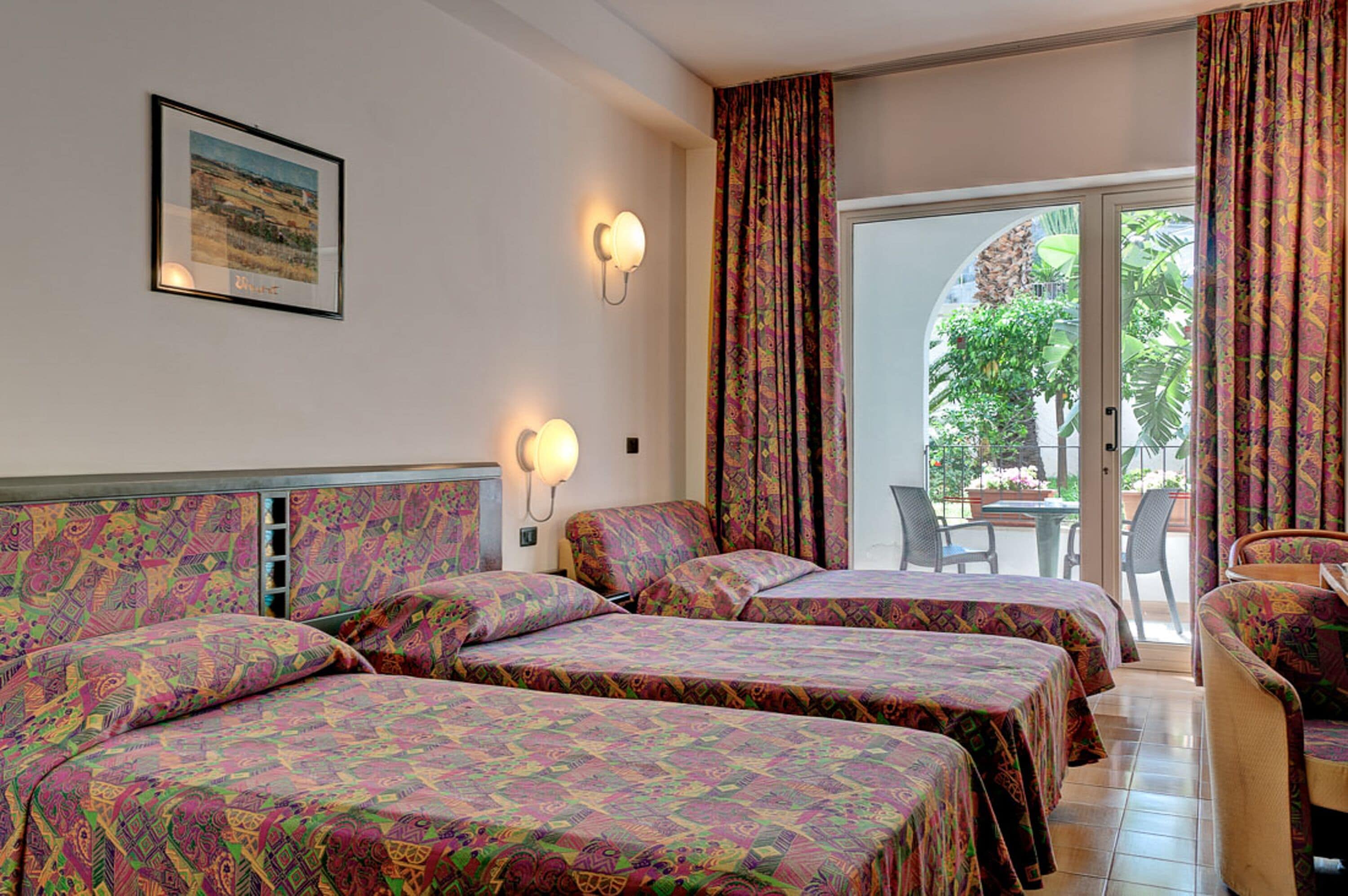 A hotel room with two beds, a dining table, and a chair. There is a window with curtains and a view of greenery outside.