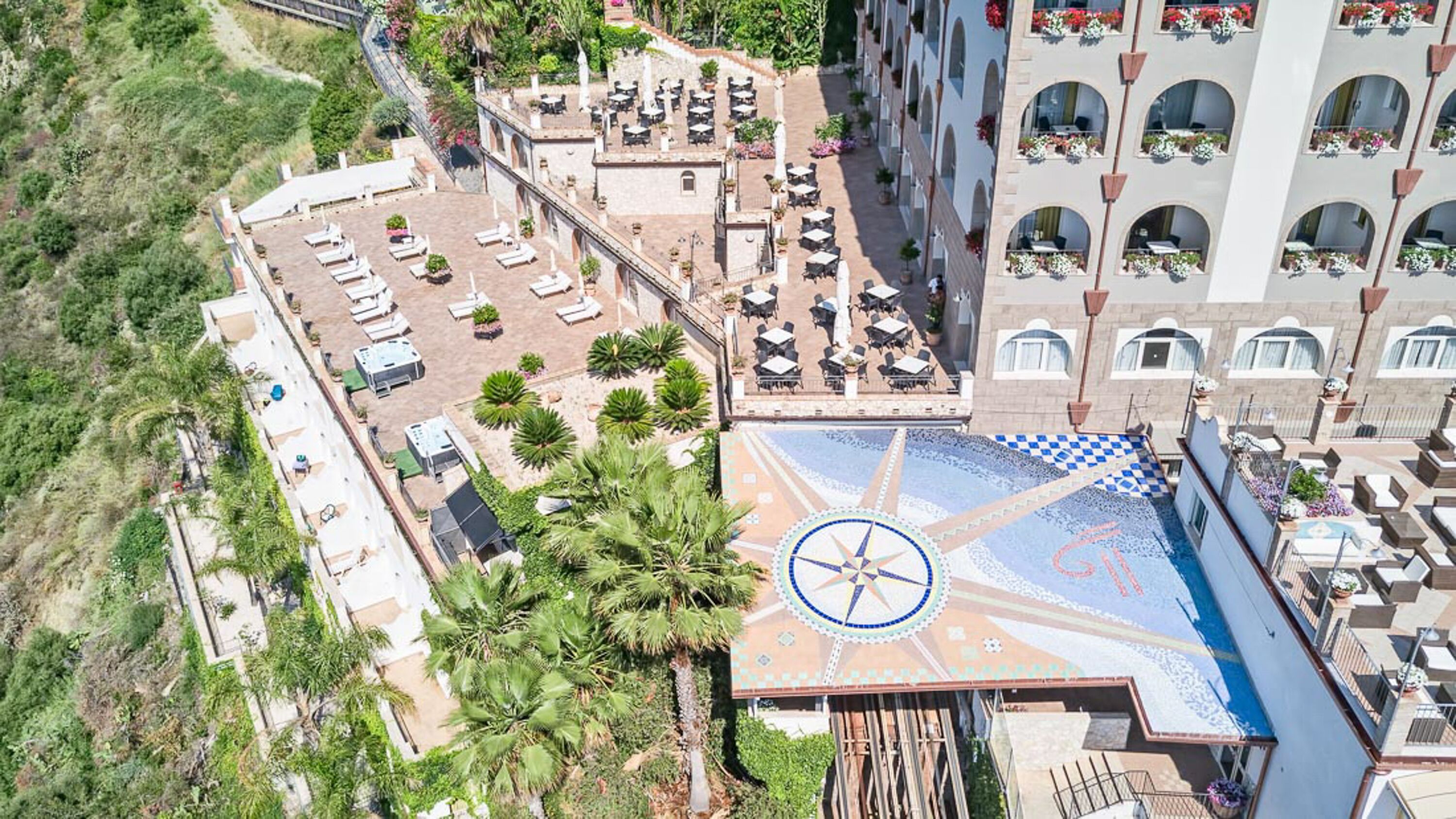 Vista aérea de un hotel con zonas de estar al aire libre, una piscina con diseño de mosaico y un edificio con balcones.