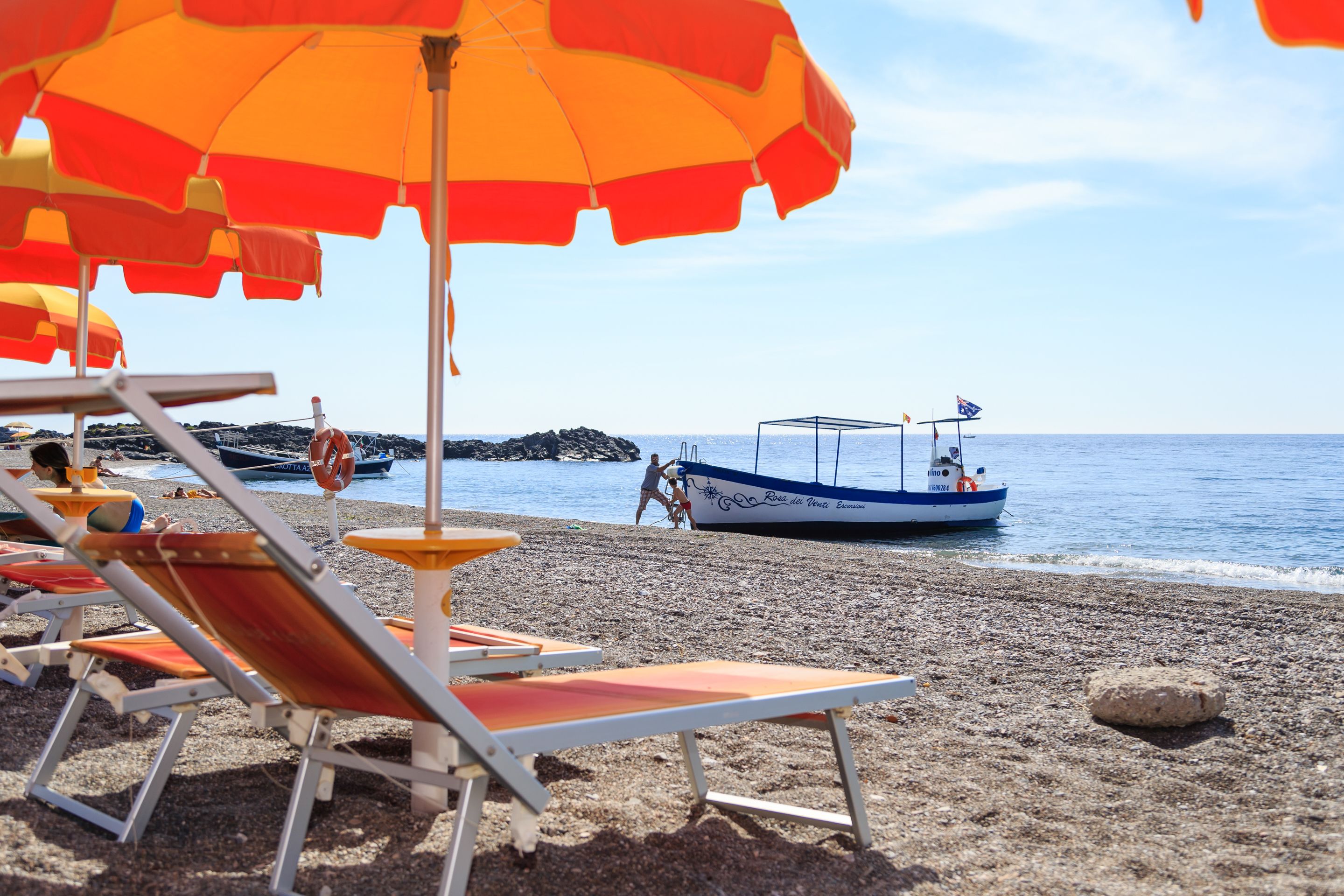 Ein Strand mit Liegestühlen und Sonnenschirmen, ein Boot sowie Felsen im Hintergrund.
