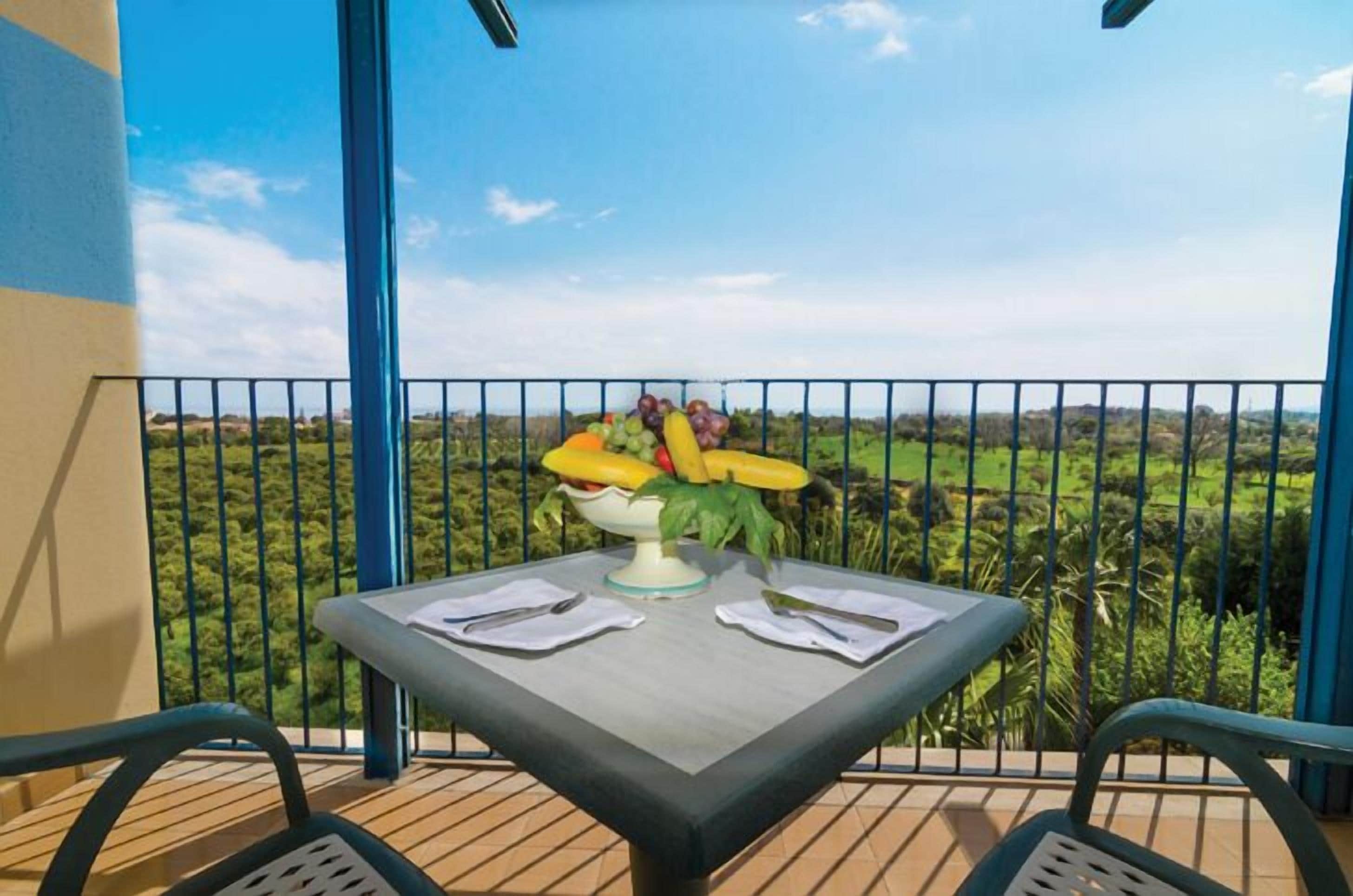 A balcony with a table set for two, featuring a fruit bowl with bananas, grapes, and other fruits.