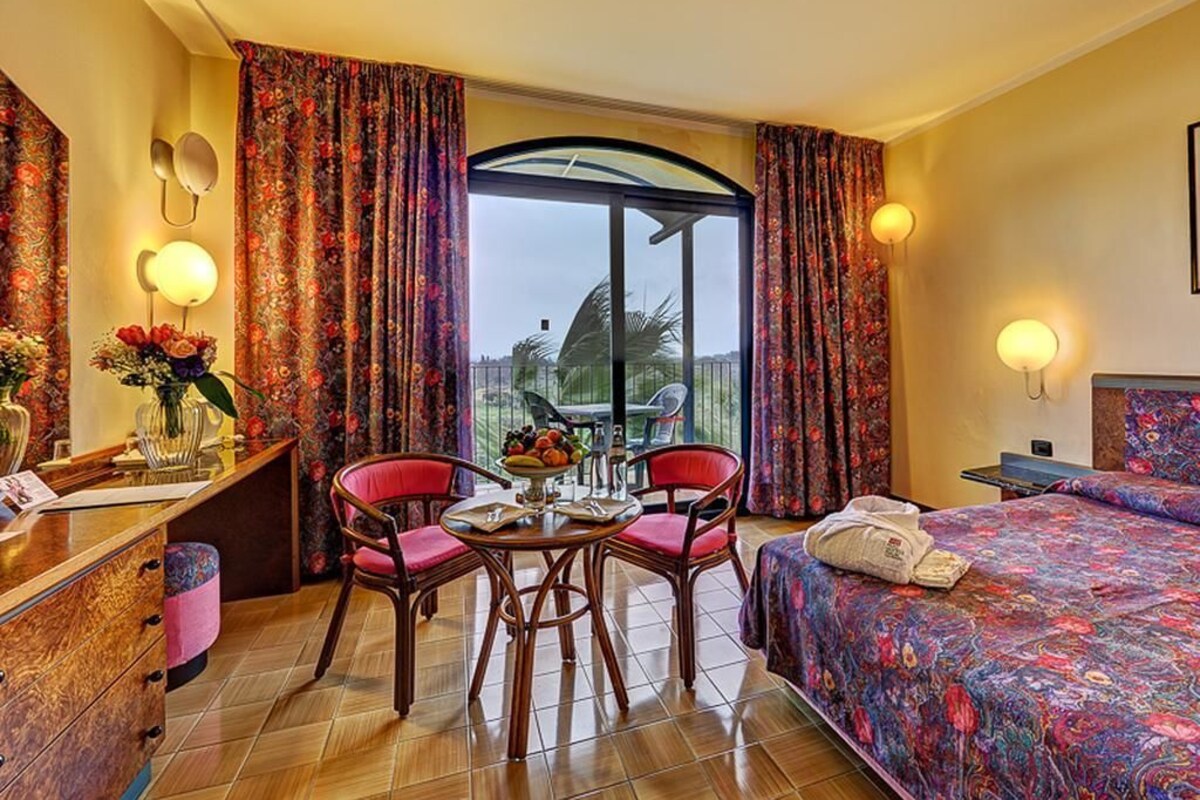 A hotel room with a bed, a desk with a chair, a table with chairs, and a window with curtains.