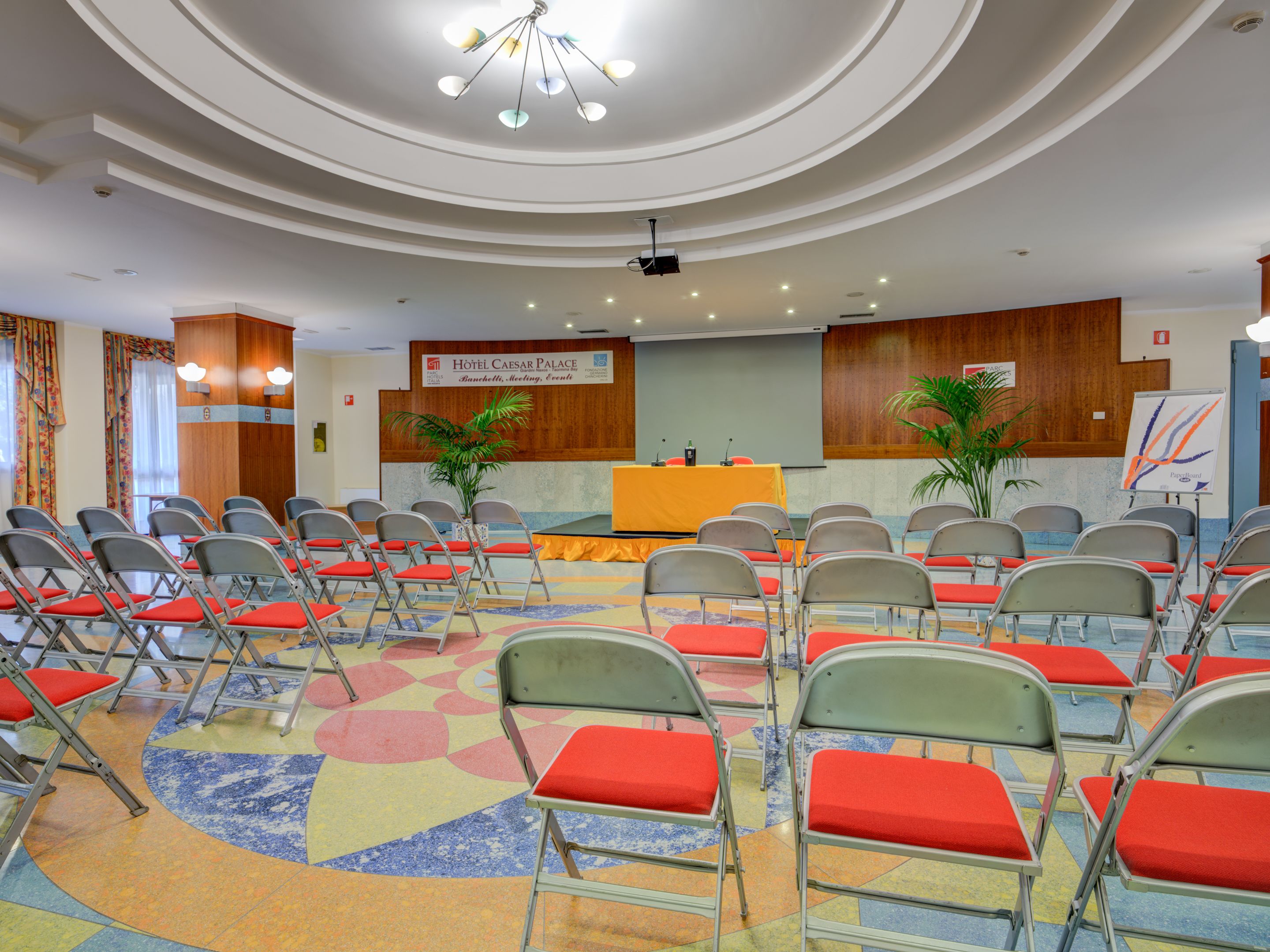 Un salón de conferencias de hotel con filas de sillas, un escenario y un cartel.