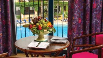 Uma mesa de jantar com um centro de frutas, uma cadeira e vista para uma piscina e edifícios.